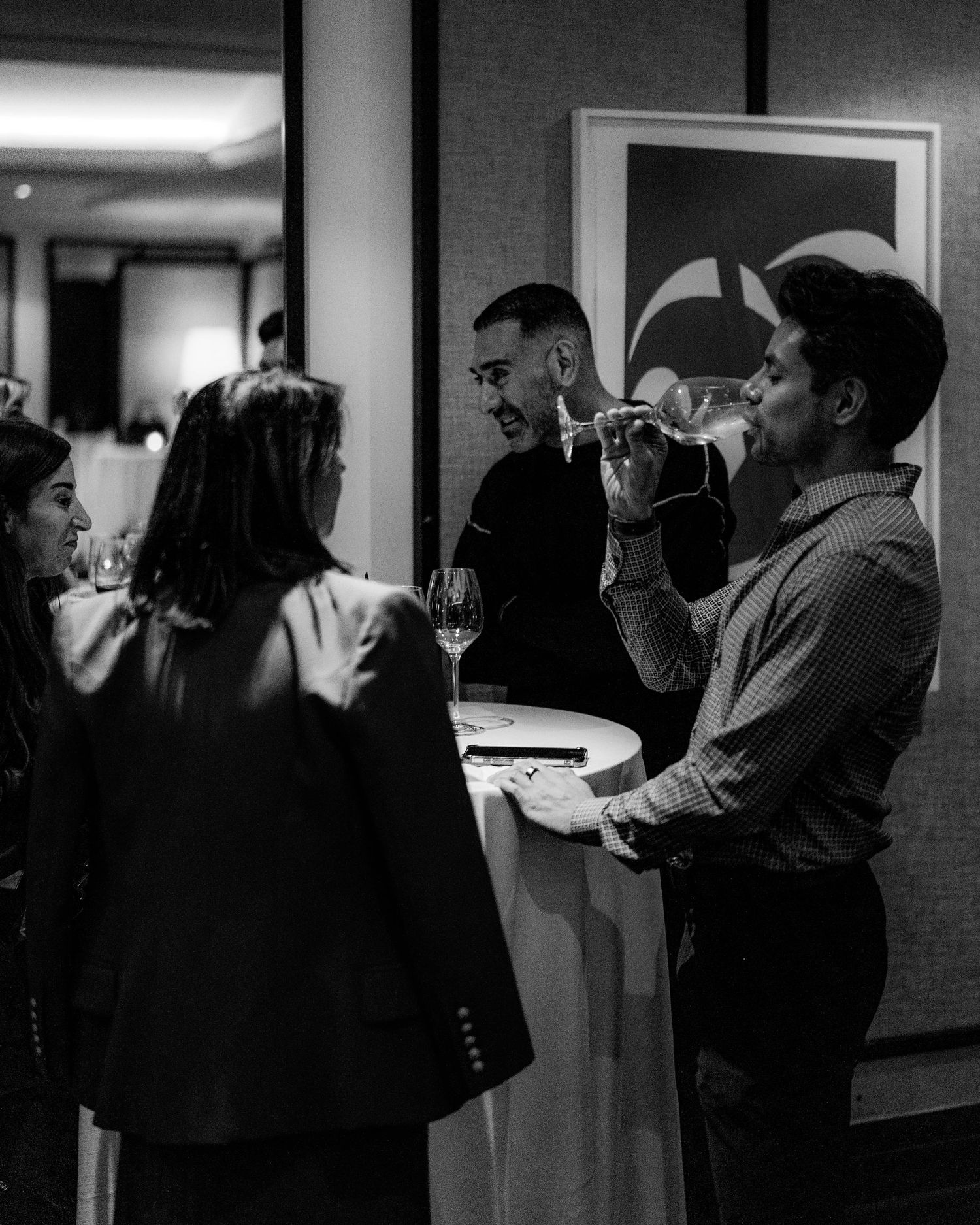 Black and white photo of guests mingling around a high-top table, holding wine glasses at an elegant evening reception.