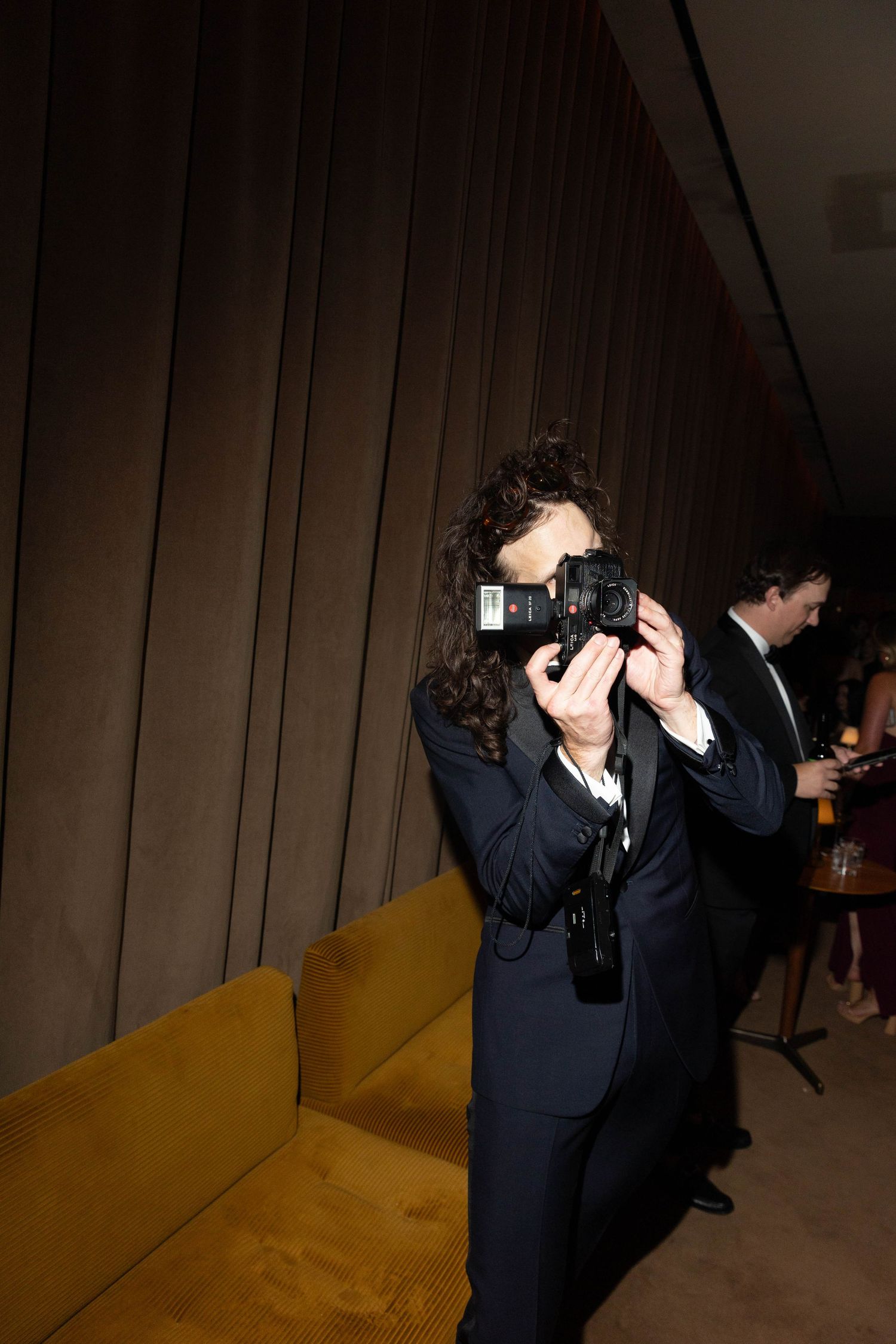 Curly-haired photographer in a dark suit aims a camera at the viewer beside a mustard velvet sofa at an indoor evening event.