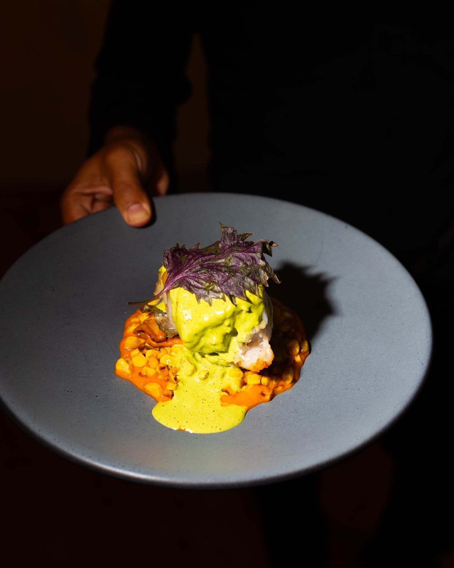 Hand holding a gray plate of gourmet fish topped with green sauce and purple microgreens over orange sauce and yellow drizzle.