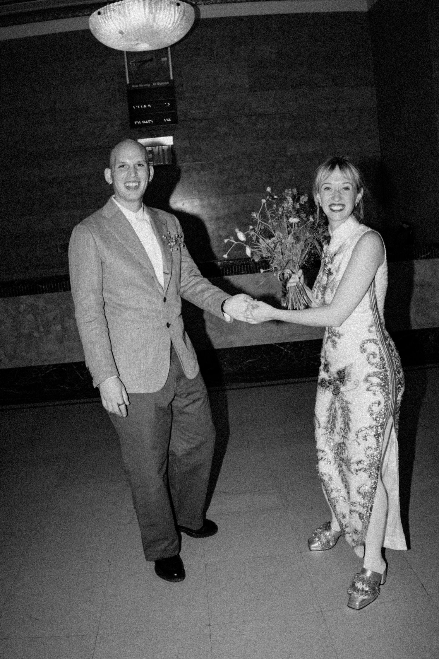 Black and white photo of bride and groom holding hands and smiling in elegant venue.