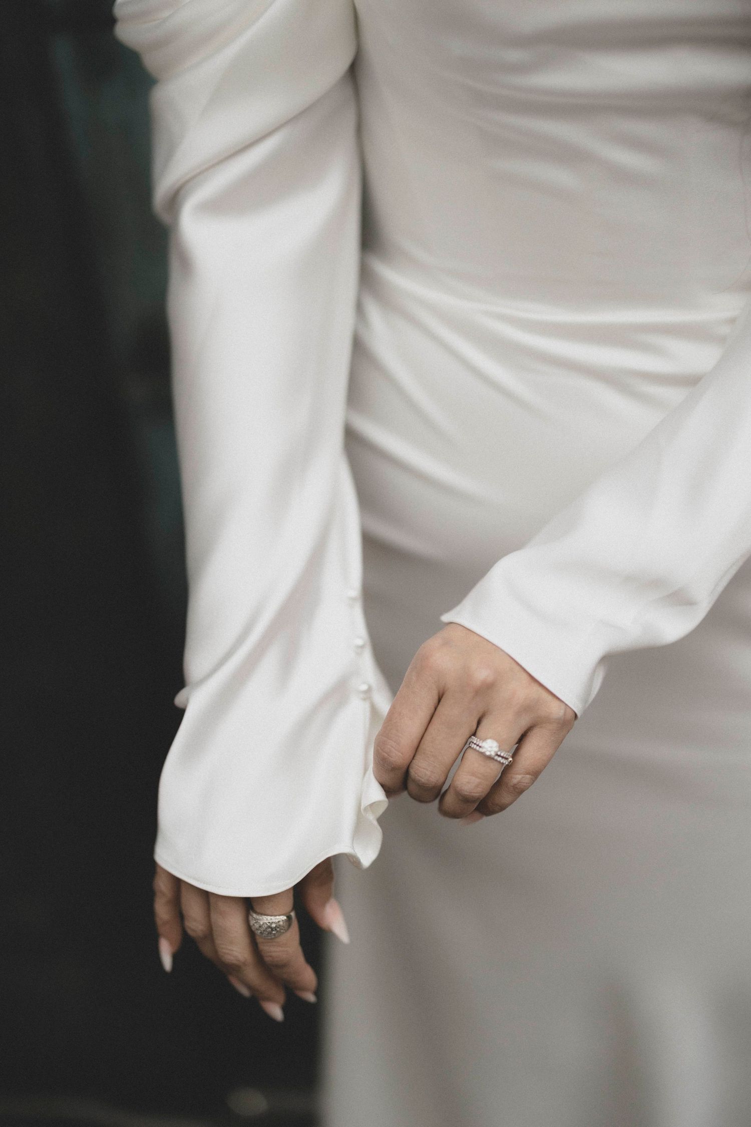 Close-up of bride's hand wearing diamond wedding ring on white satin long-sleeve wedding dress.