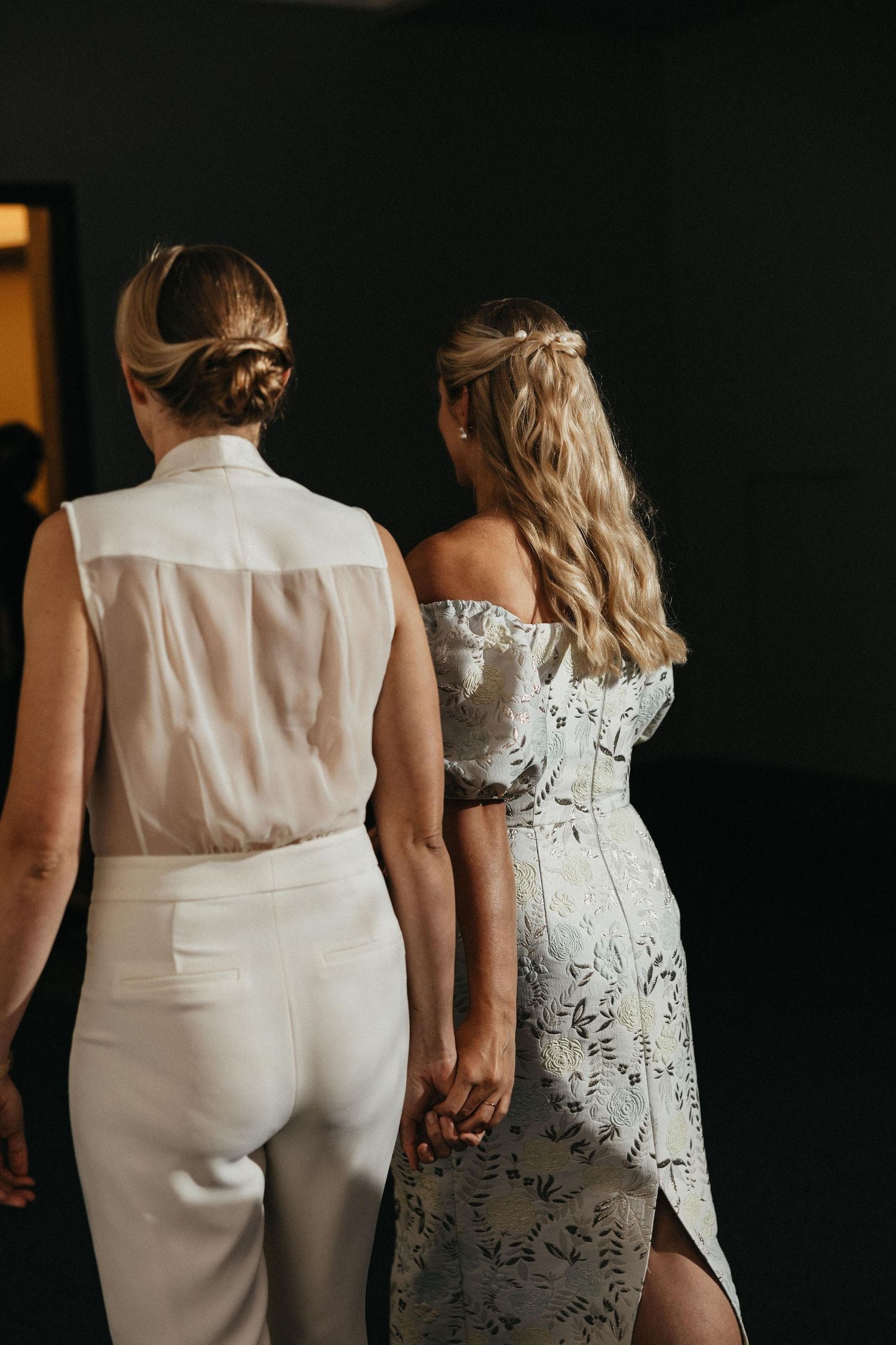 Rear view of two brides holding hands walking together in white wedding attire.