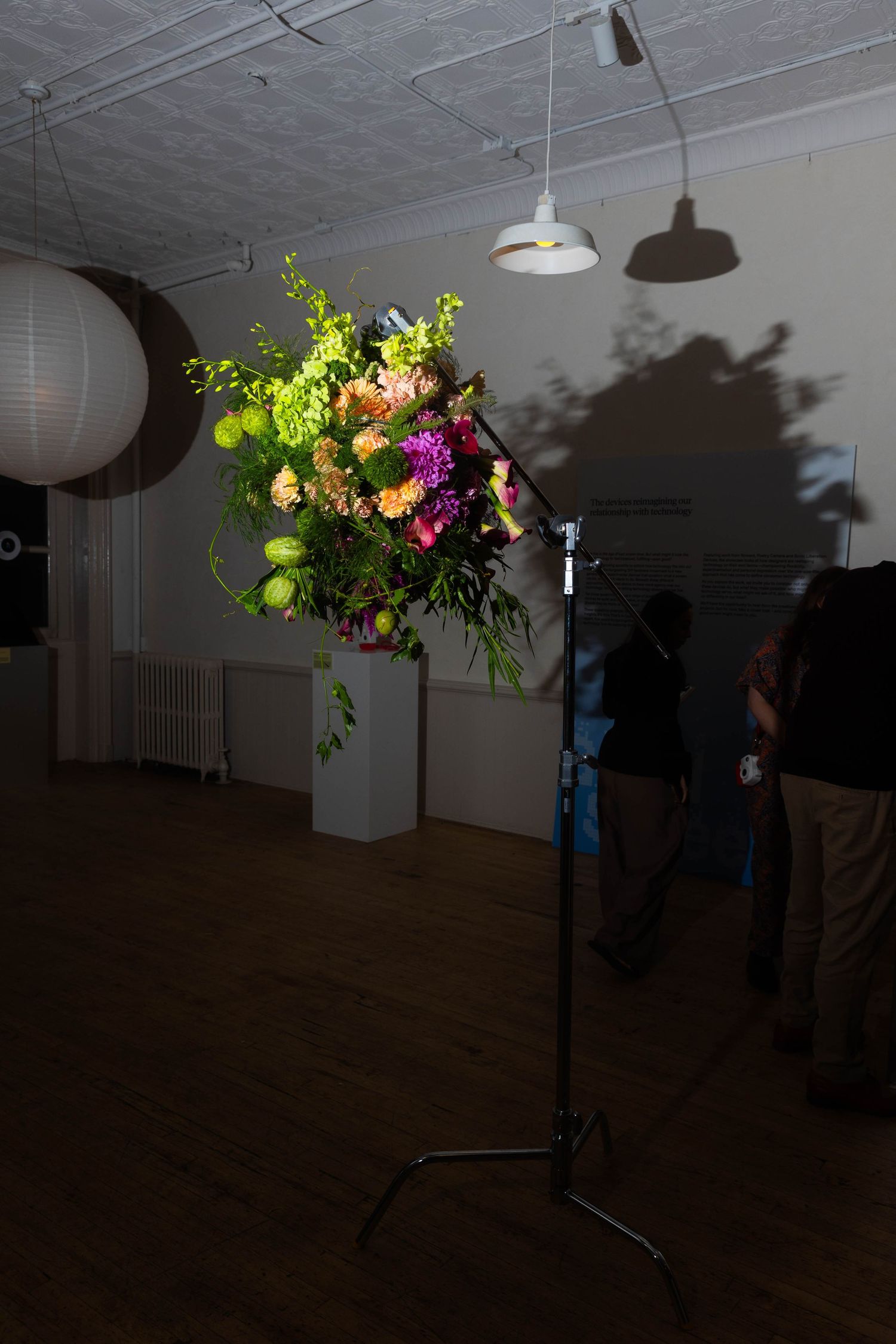 Colorful floral arrangement mounted on a light stand in a dim loft gallery, casting shadows on the wall as visitors stand nearby.