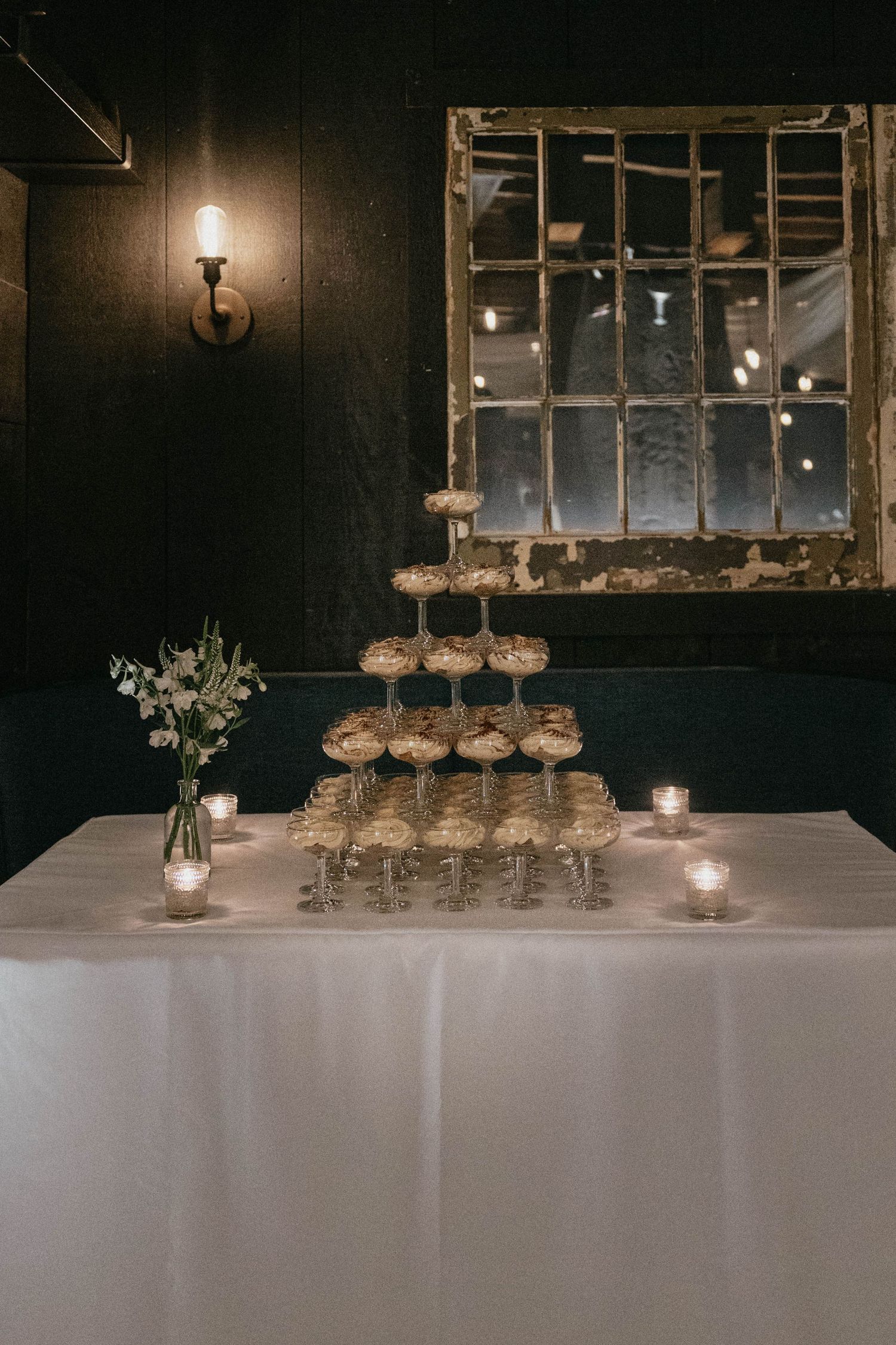 Champagne coupe tower on a white-draped table with candles and a white flower vase, set against a rustic vintage window mirror.
