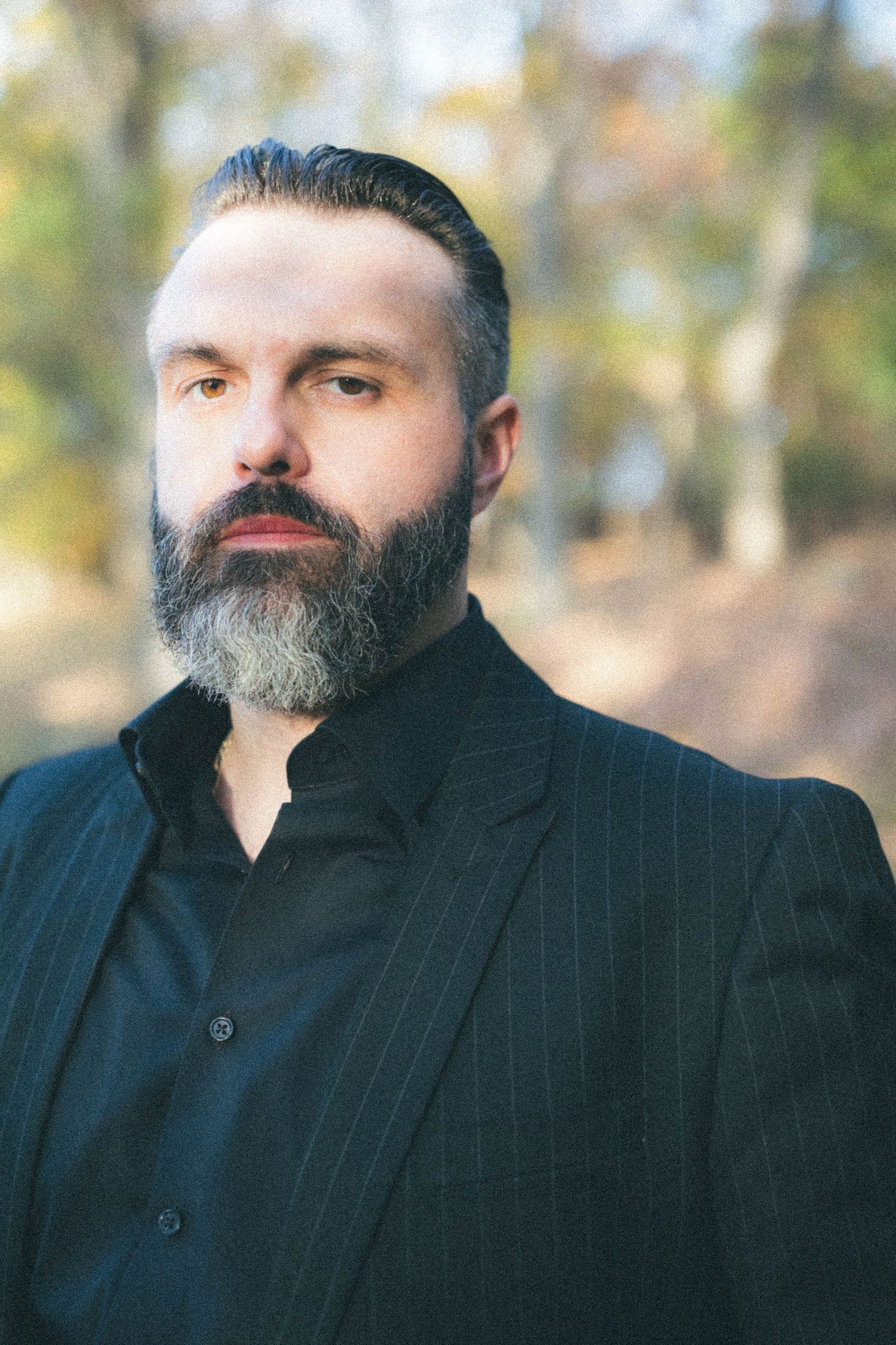 Bearded man in a black pinstripe suit posing outdoors with autumn trees in background.