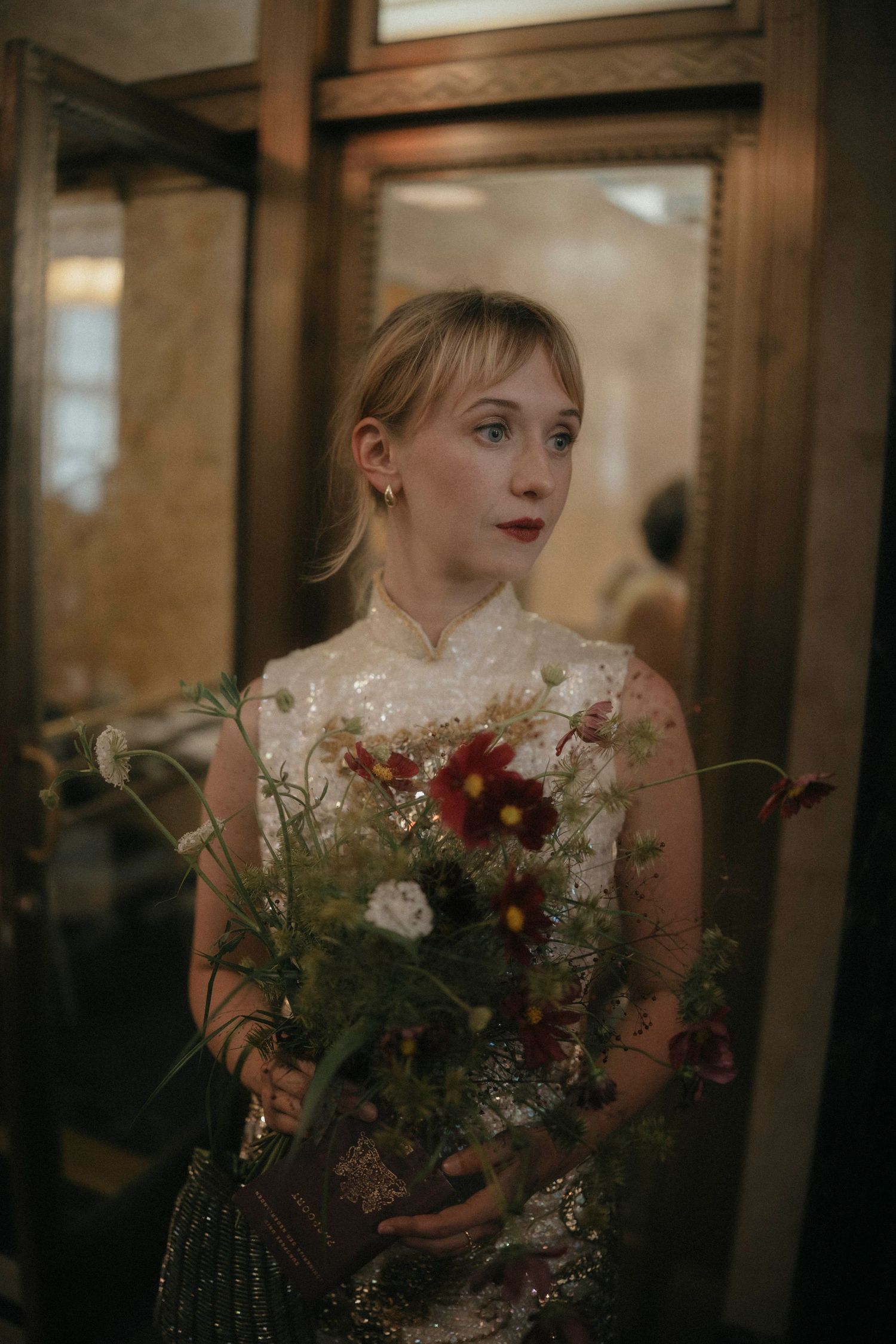 Bride in sequined gown holds wildflower bouquet with red and white blooms, standing by ornate mirrors.