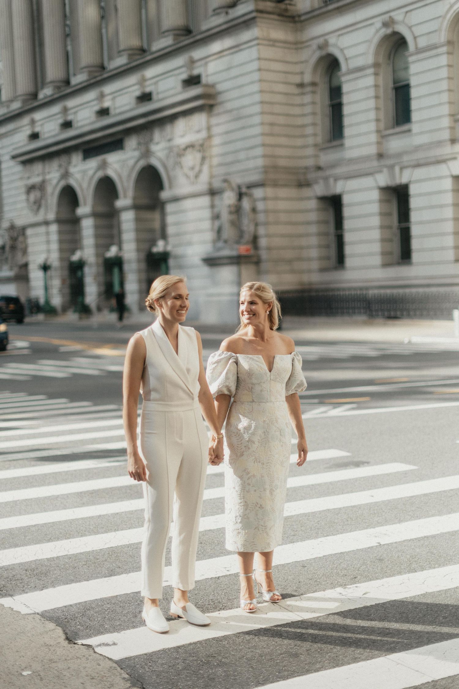 Two brides in white holding hands while crossing a sunny city street on their wedding day.