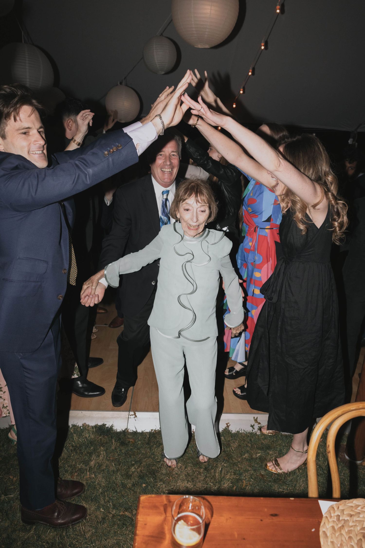 Elderly woman in mint suit walks under tunnel of raised hands at outdoor wedding celebration.