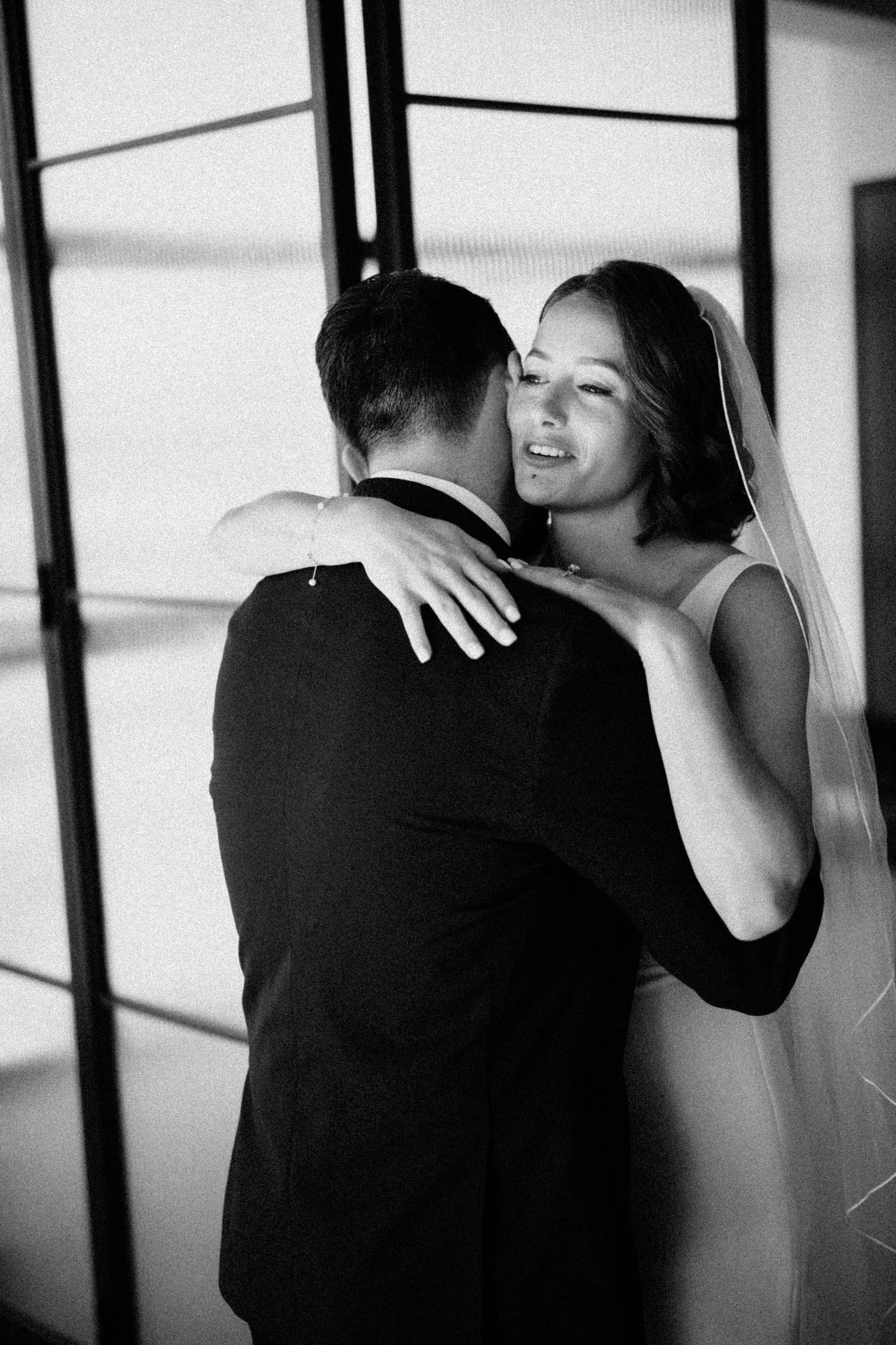 Black and white photo of smiling bride embracing groom near large windows at their wedding.