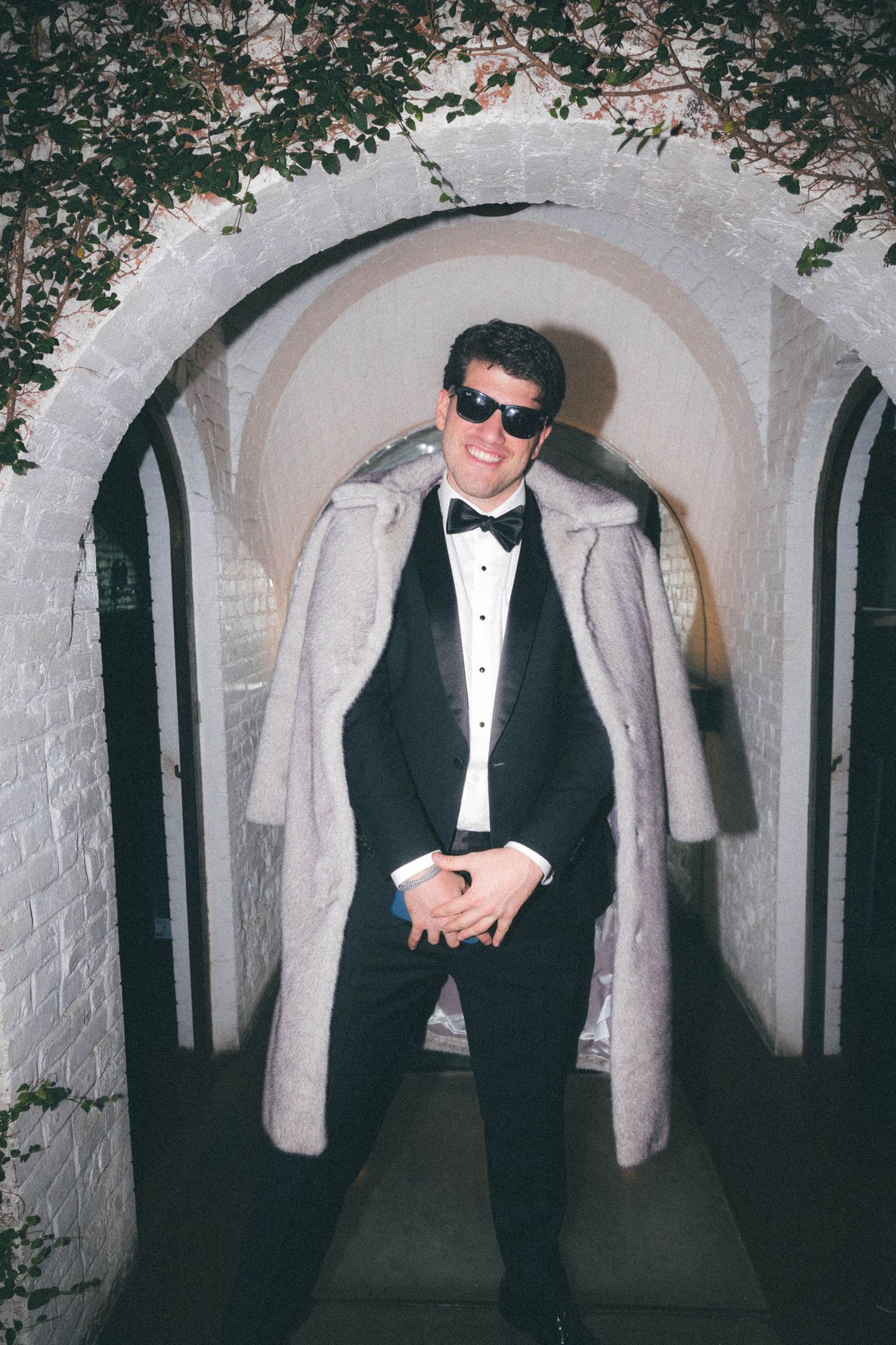 A stylish man in a tuxedo and fur coat poses under stone arches with sunglasses at night.