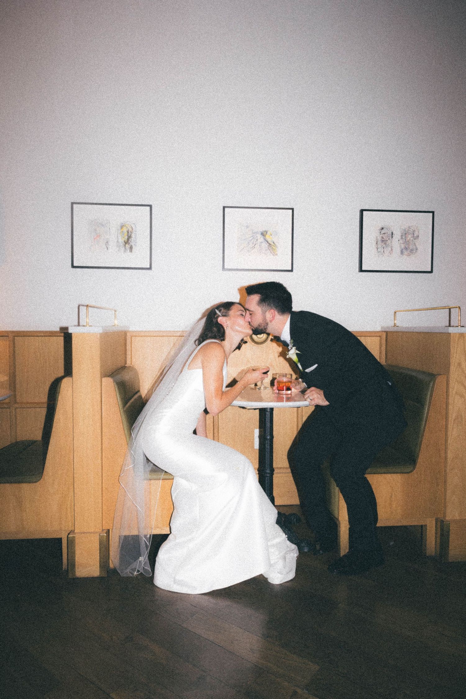 Bride and groom share a kiss over a small cake at a wooden booth inside their wedding venue.