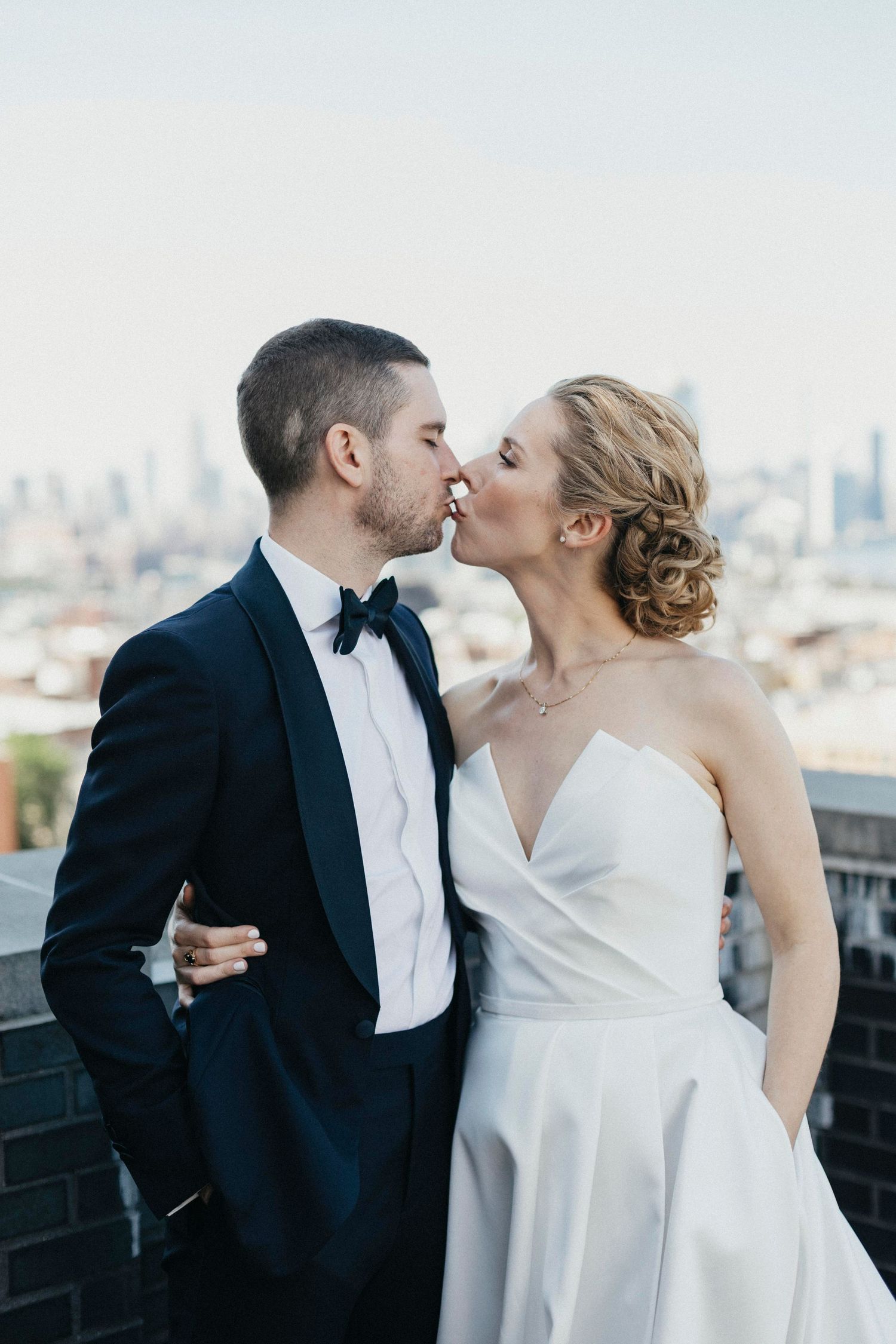 Bride and groom sharing a romantic kiss on rooftop with city skyline backdrop, groom in navy tuxedo.