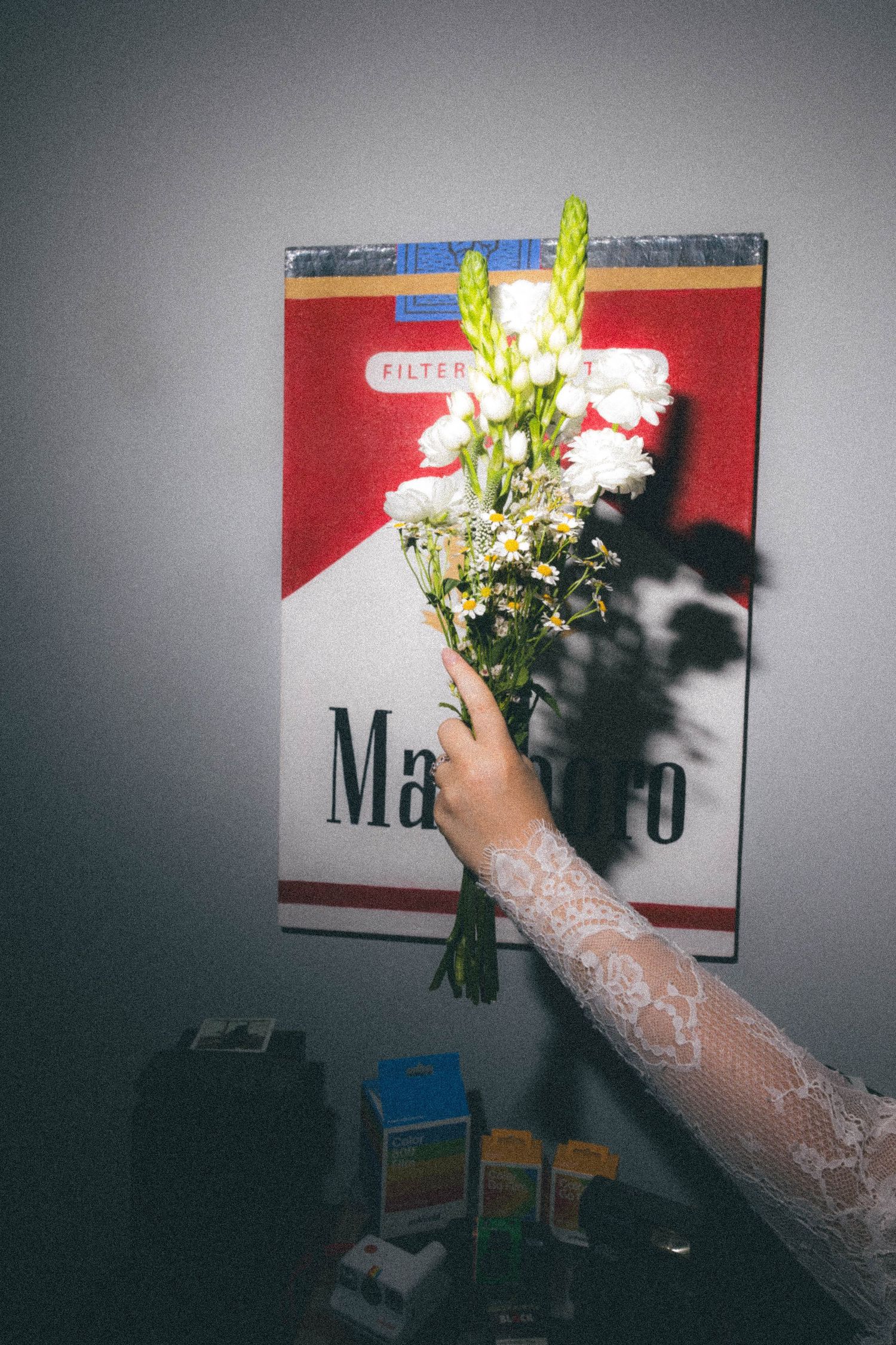 Hand holding white flowers against a large Marlboro cigarette pack poster on a gray wall.