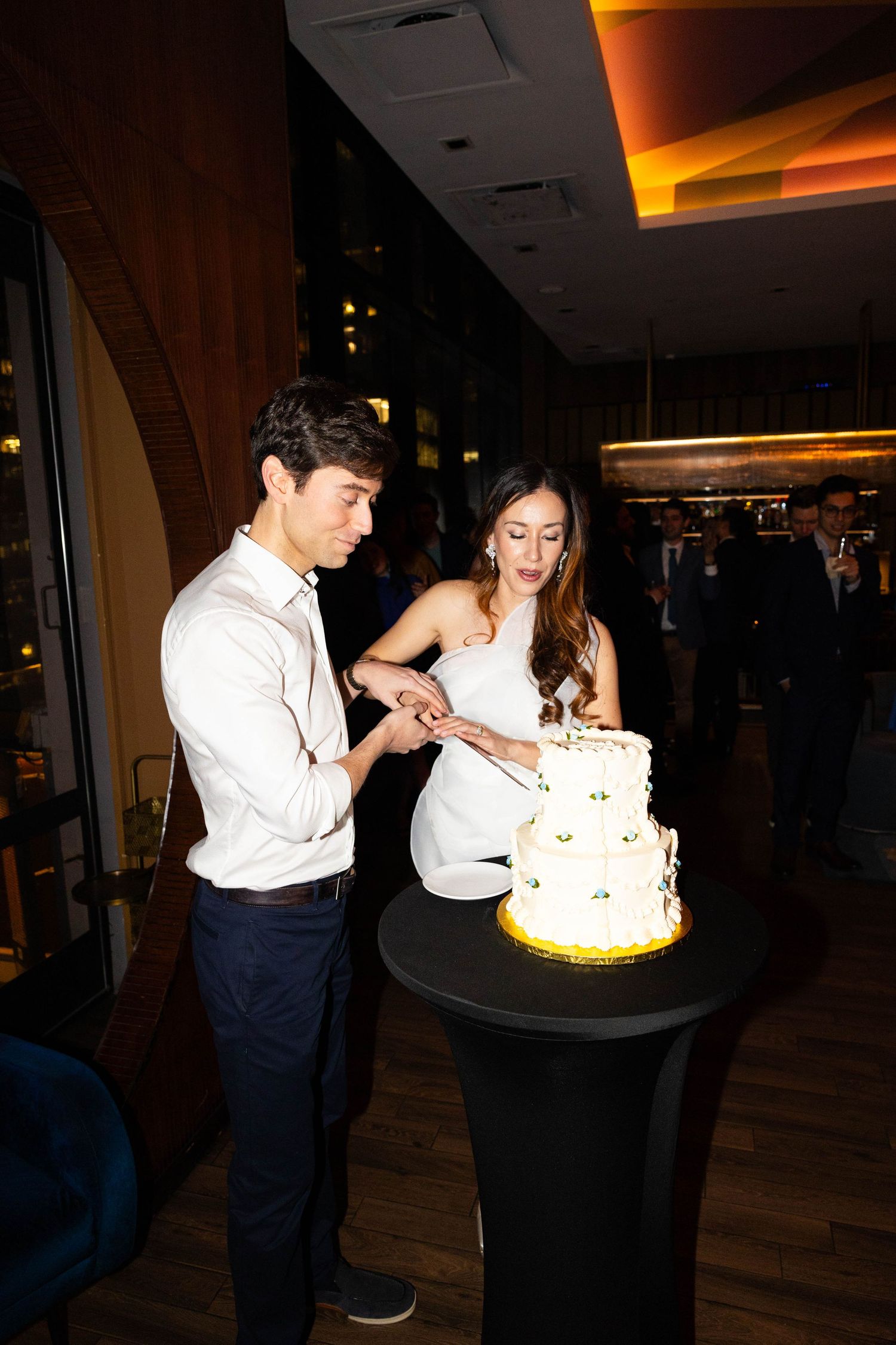 Couple in white outfits cutting three-tier white wedding cake together at elegant evening reception.