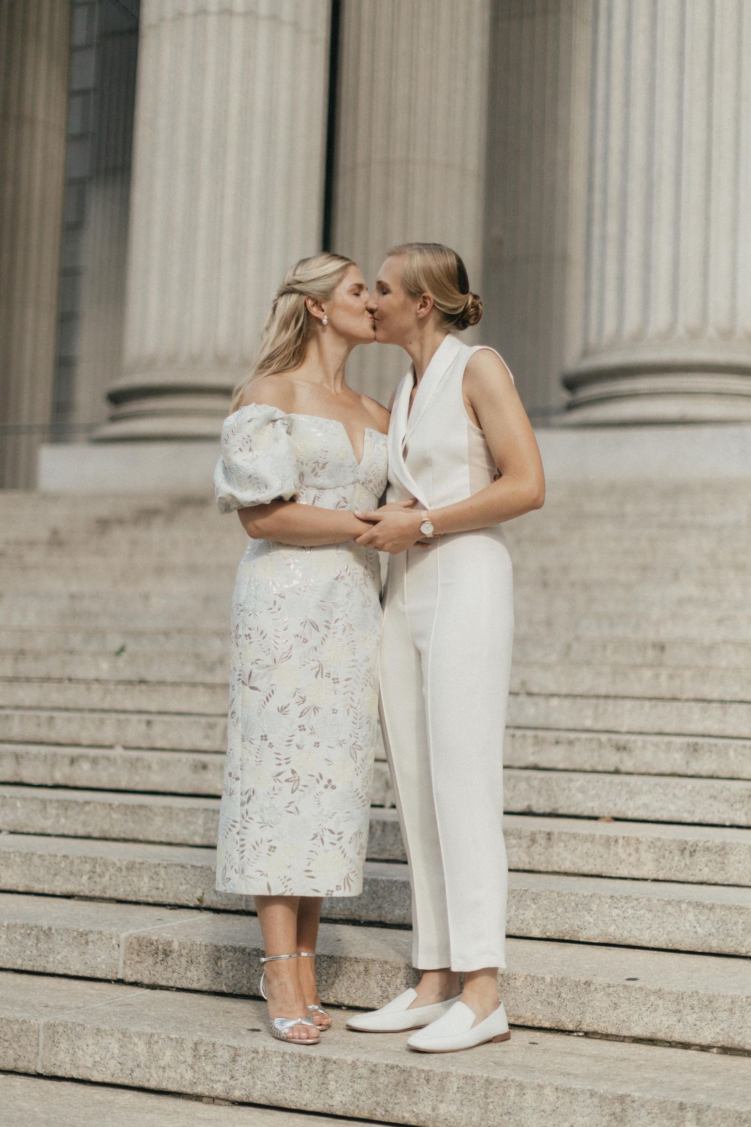 Two brides share a kiss on stone steps, one in floral dress, one in white suit.