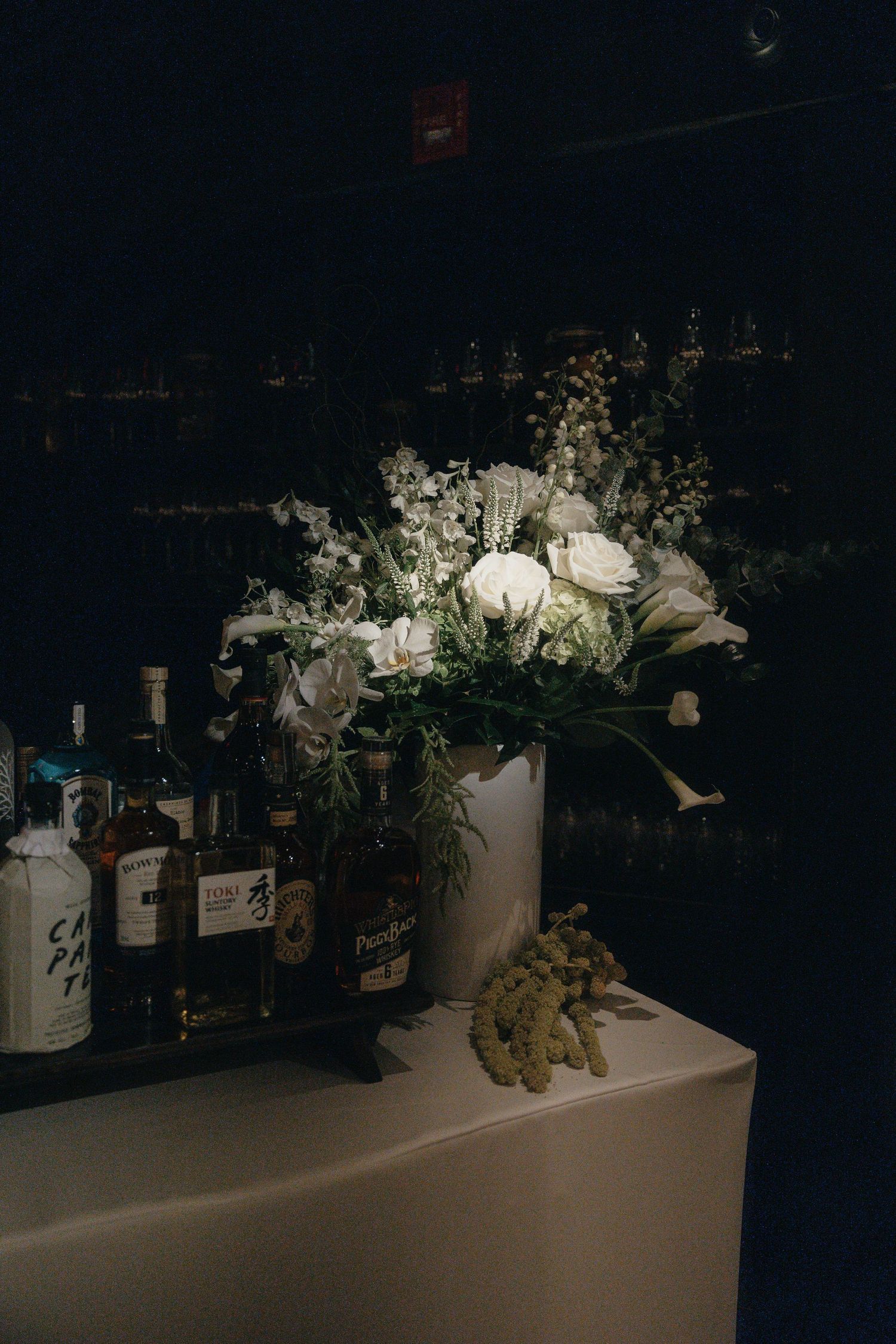 White rose and floral arrangement in a tall vase on a draped bar table beside bottles of whisky and liquor in a dimly lit bar.