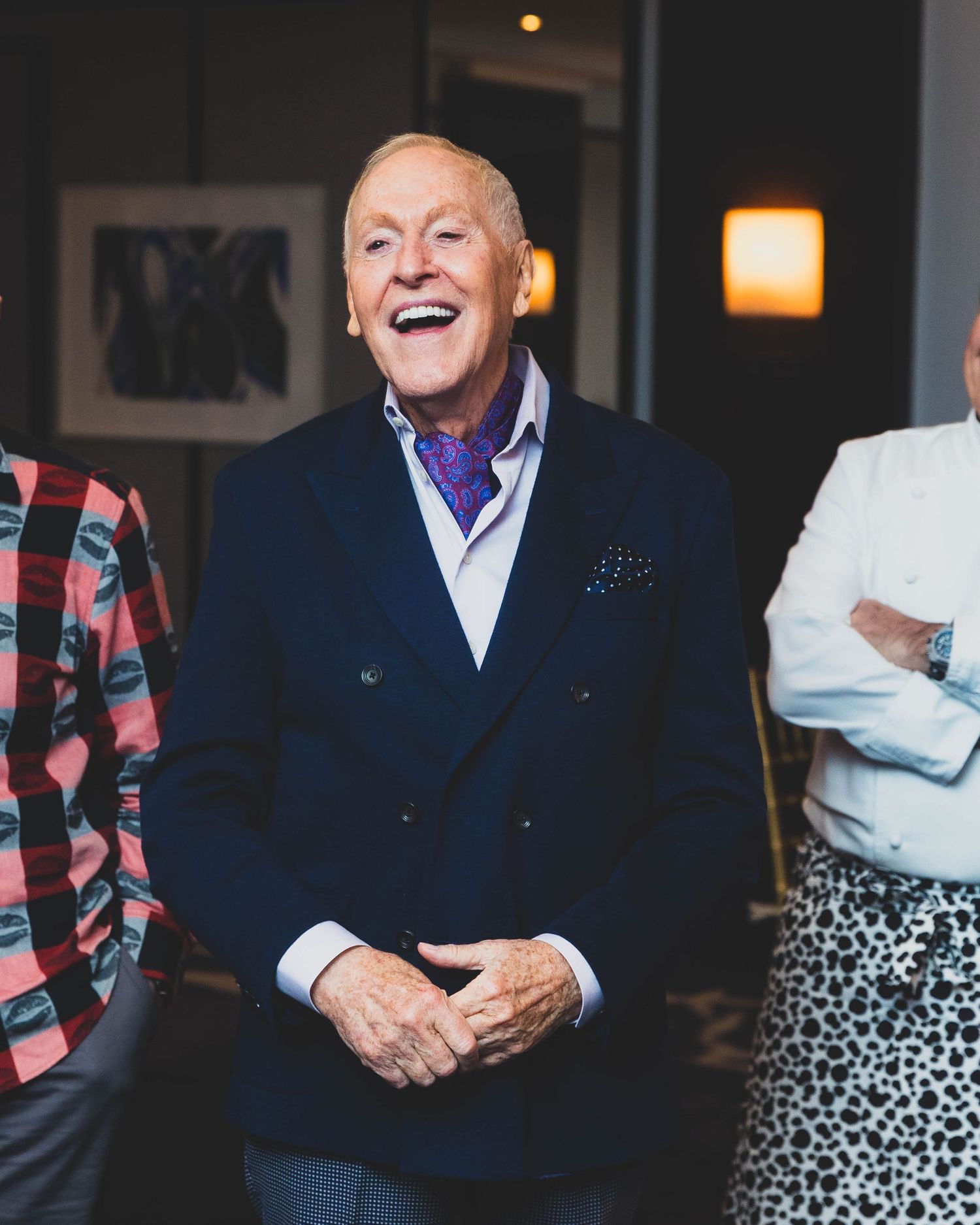 Smiling older man in a navy double-breasted blazer and purple paisley ascot laughs warmly beside two people in a softly lit room.