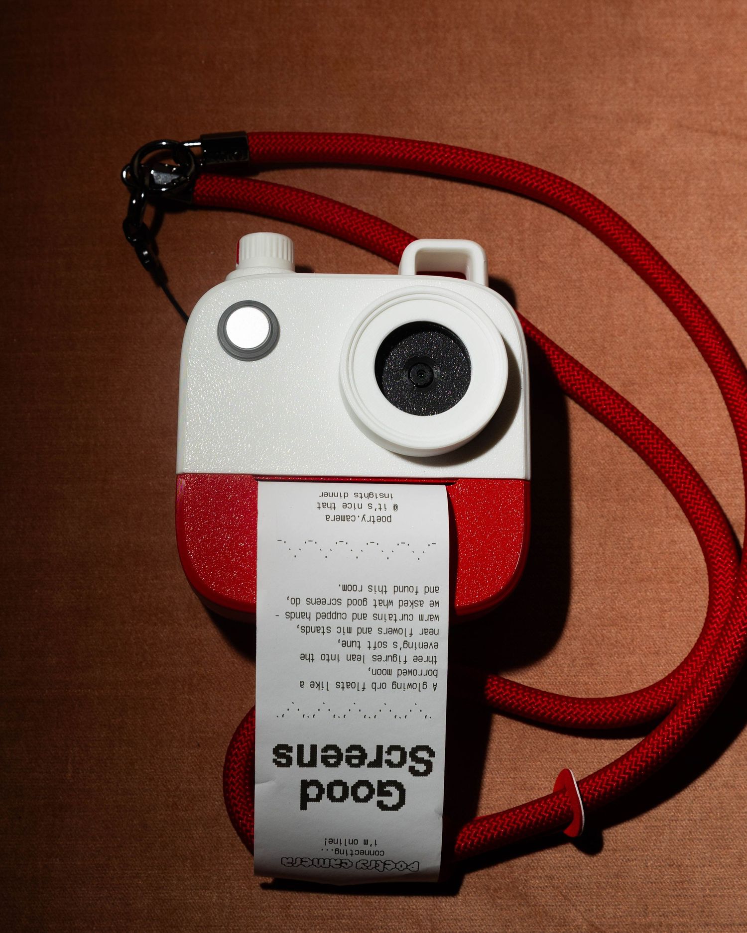 White and red toy-style camera on a red cord prints a paper receipt reading Good Screens against a rust-colored background.