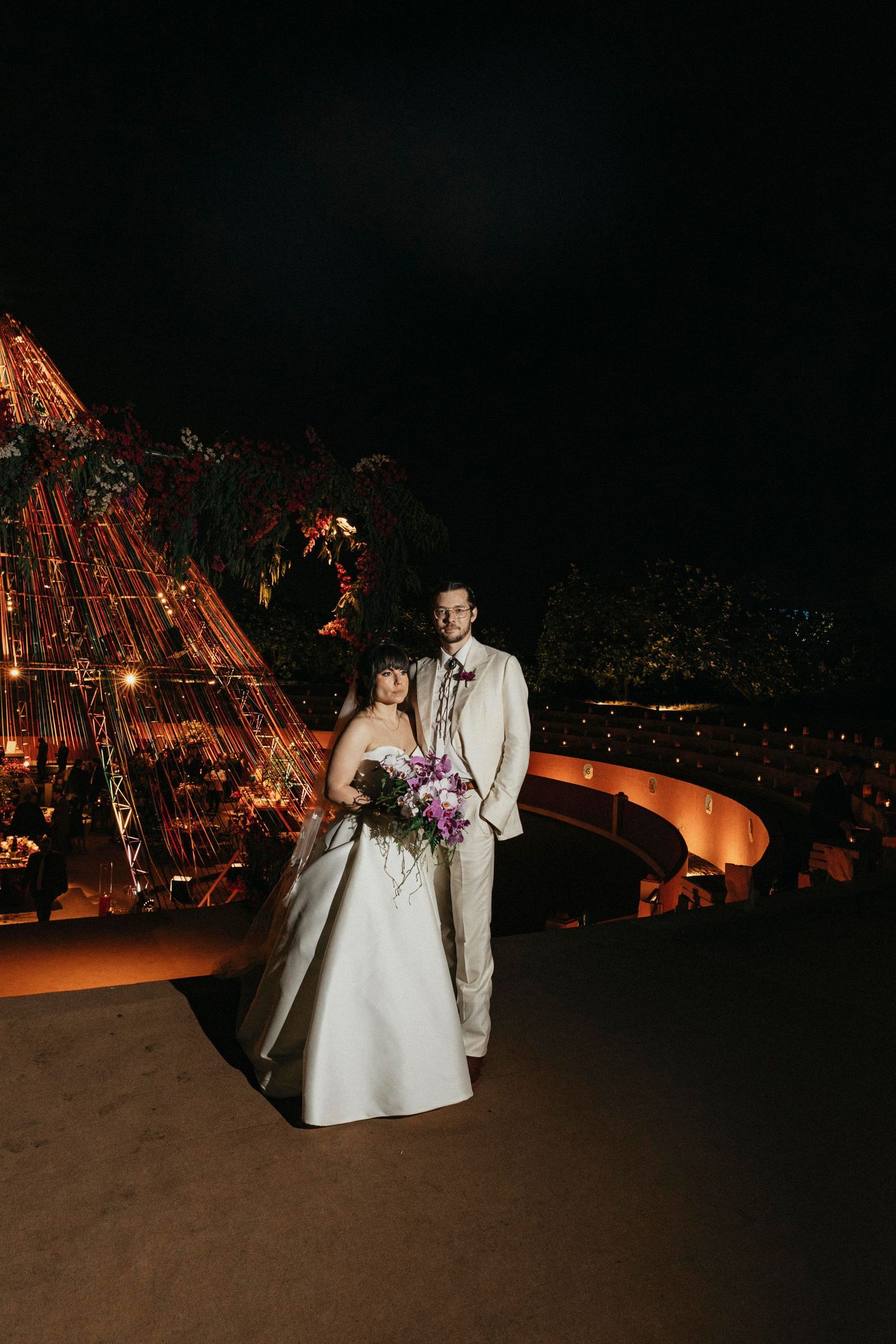 Bride and groom pose elegantly at night beside a stunning illuminated teepee wedding reception venue.