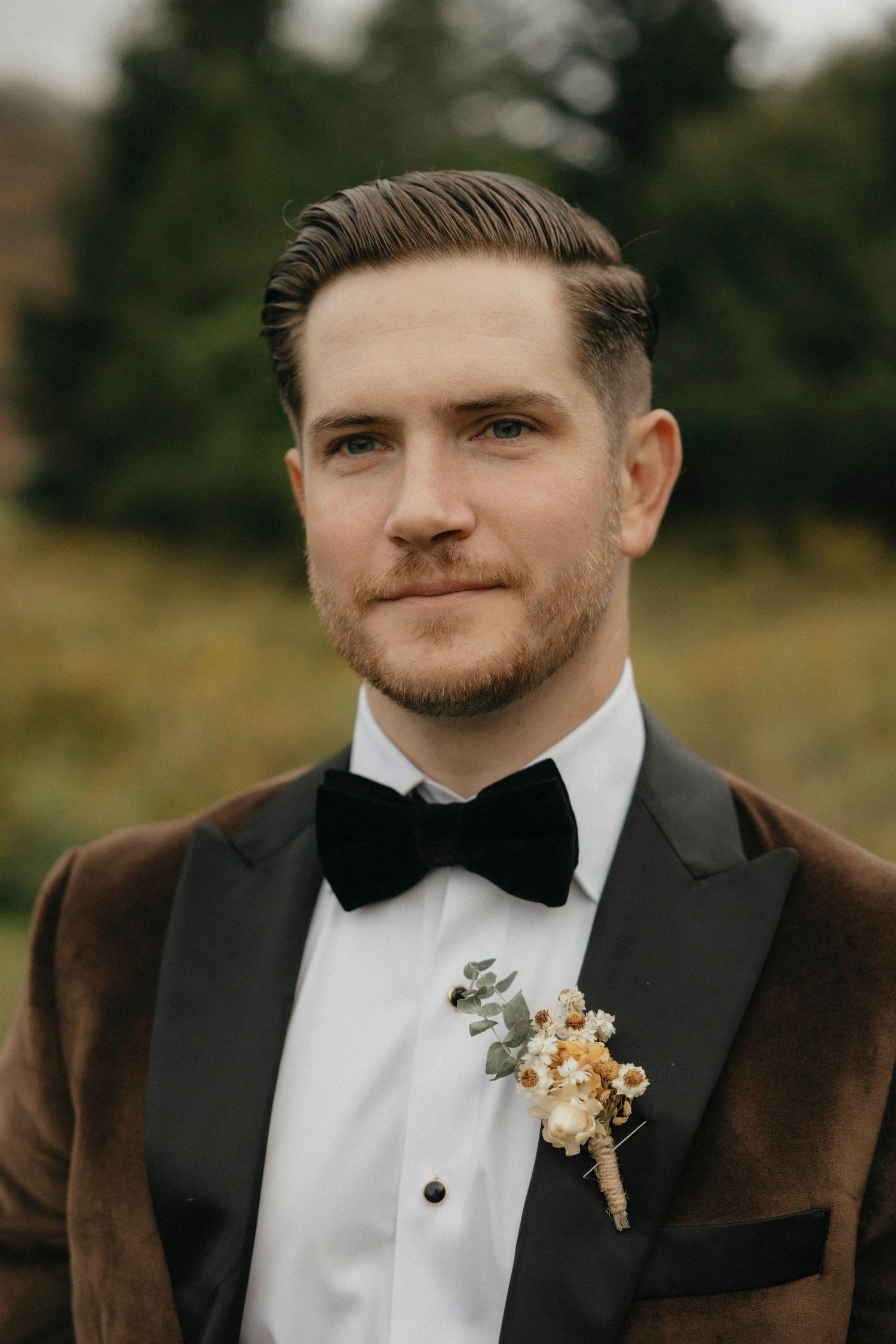 Handsome groom in brown velvet tuxedo with black bow tie and floral boutonniere outdoors.
