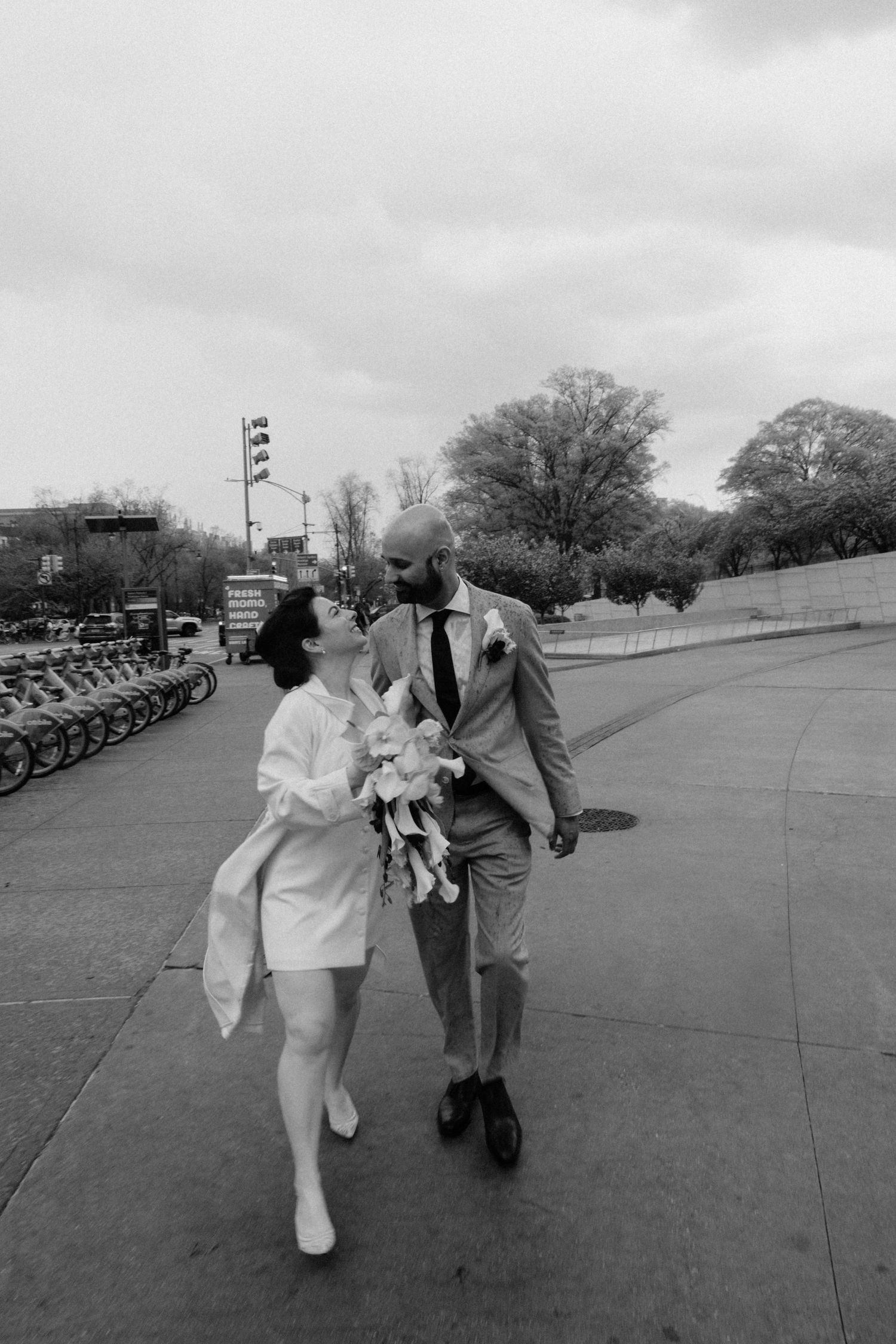 Bride and groom walk together outdoors in black and white, smiling at each other on sidewalk.