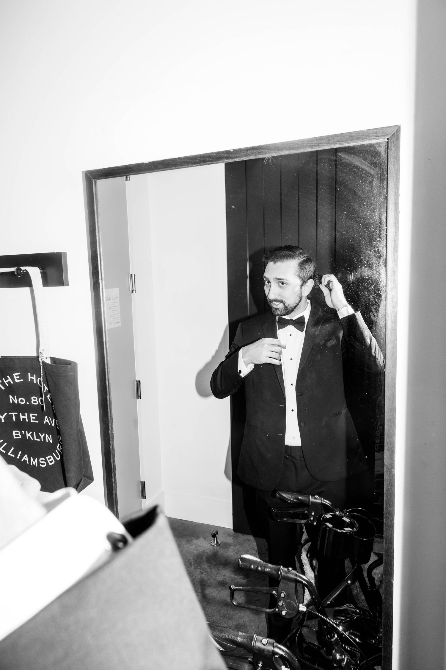 Groom in tuxedo adjusting bow tie while looking in mirror reflection in black and white wedding photo.