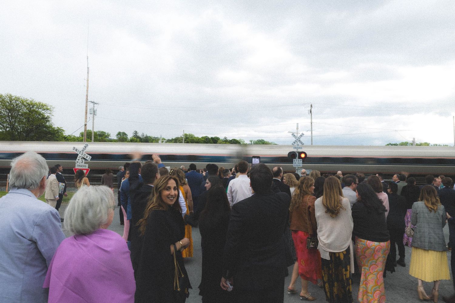 Wedding guests gathered on a train platform watching a passenger train pass by on a cloudy day.