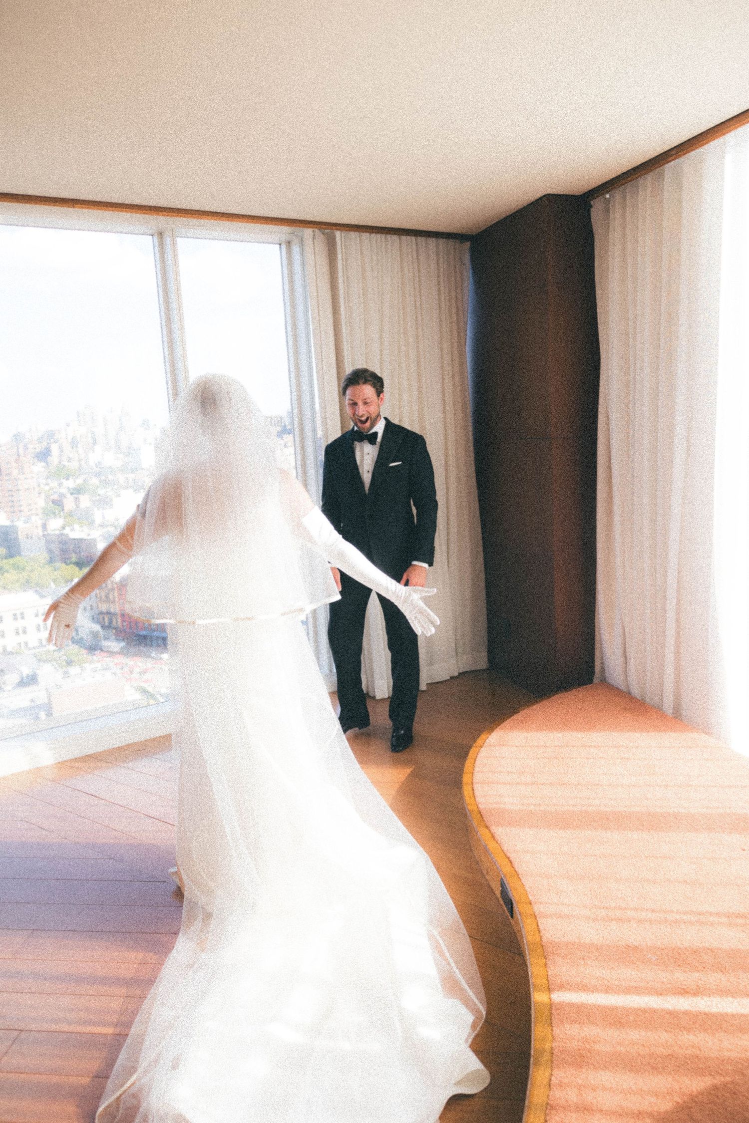 Groom in black tuxedo smiles at bride in white wedding gown and veil during first look in hotel room.