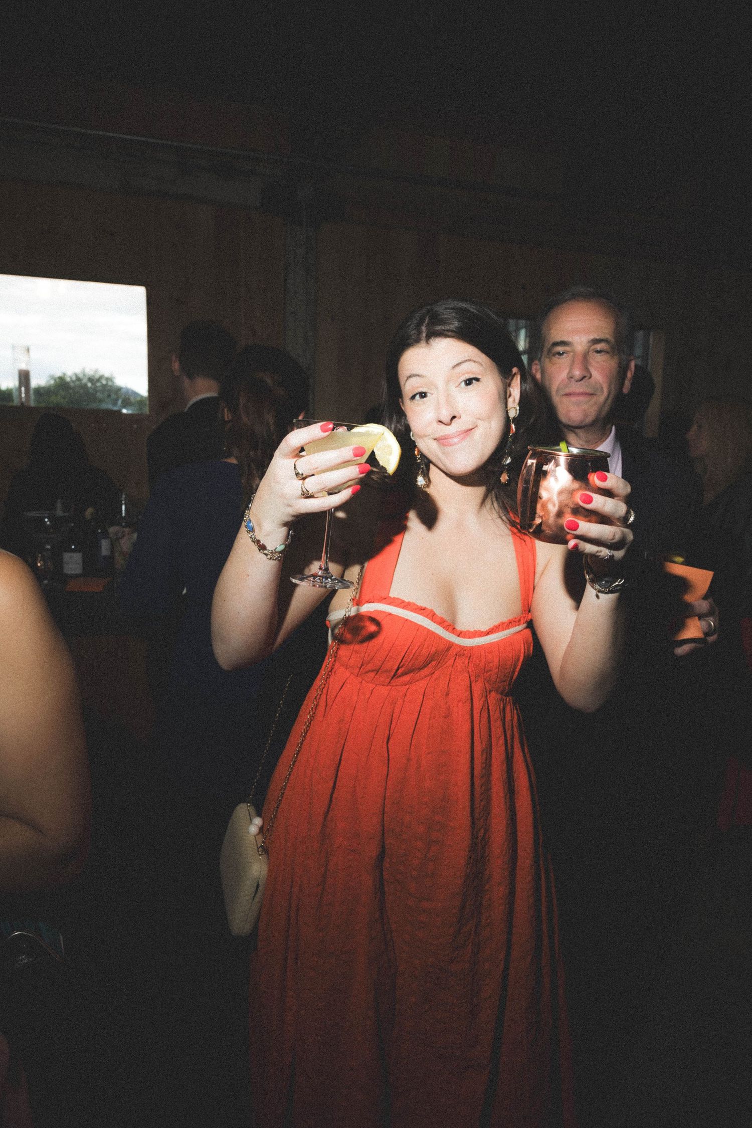 Smiling woman in red dress raising a martini glass at a lively wedding reception party.