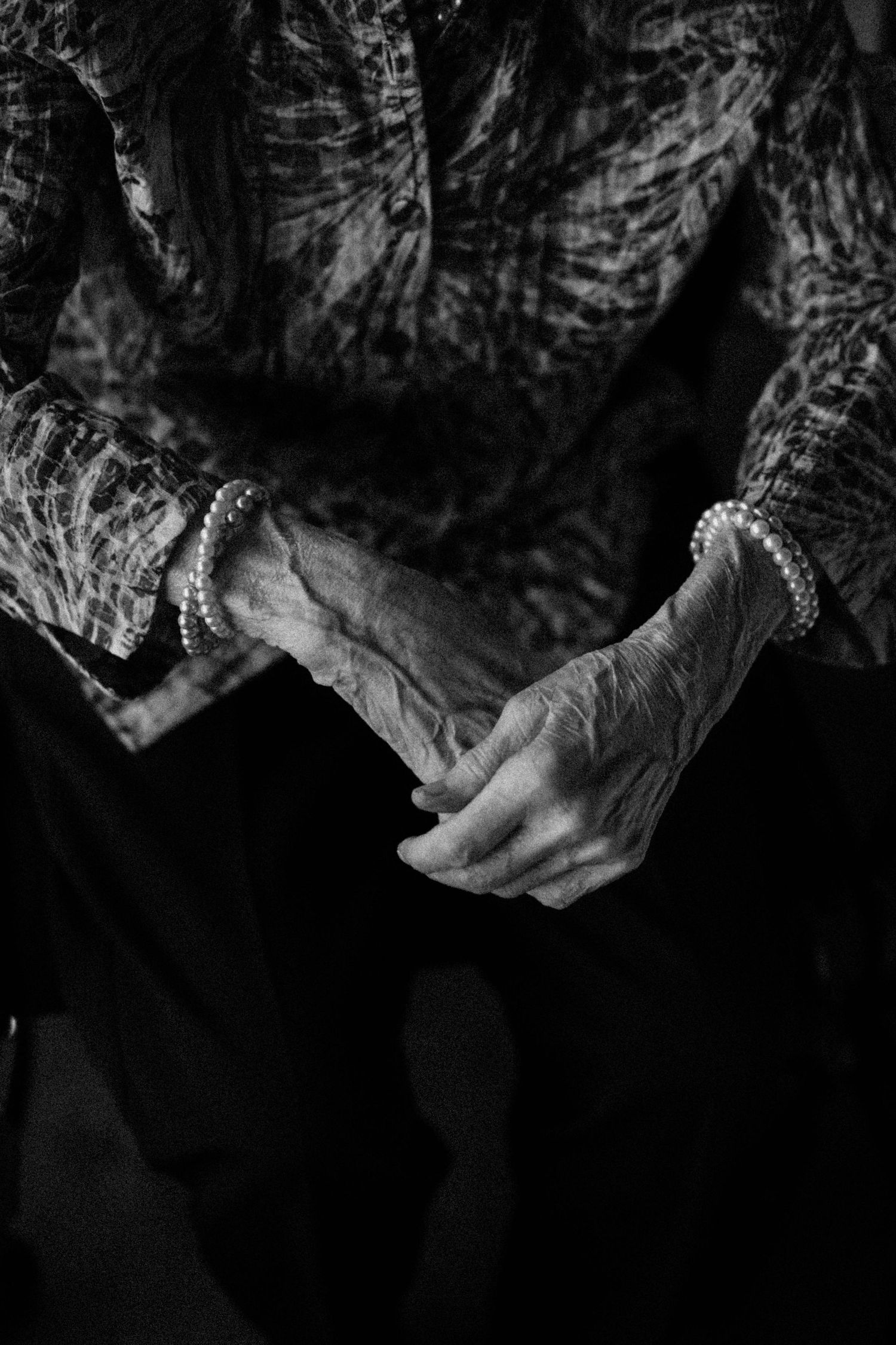 Black and white close-up of elderly womans folded hands wearing pearl bracelets at a wedding.