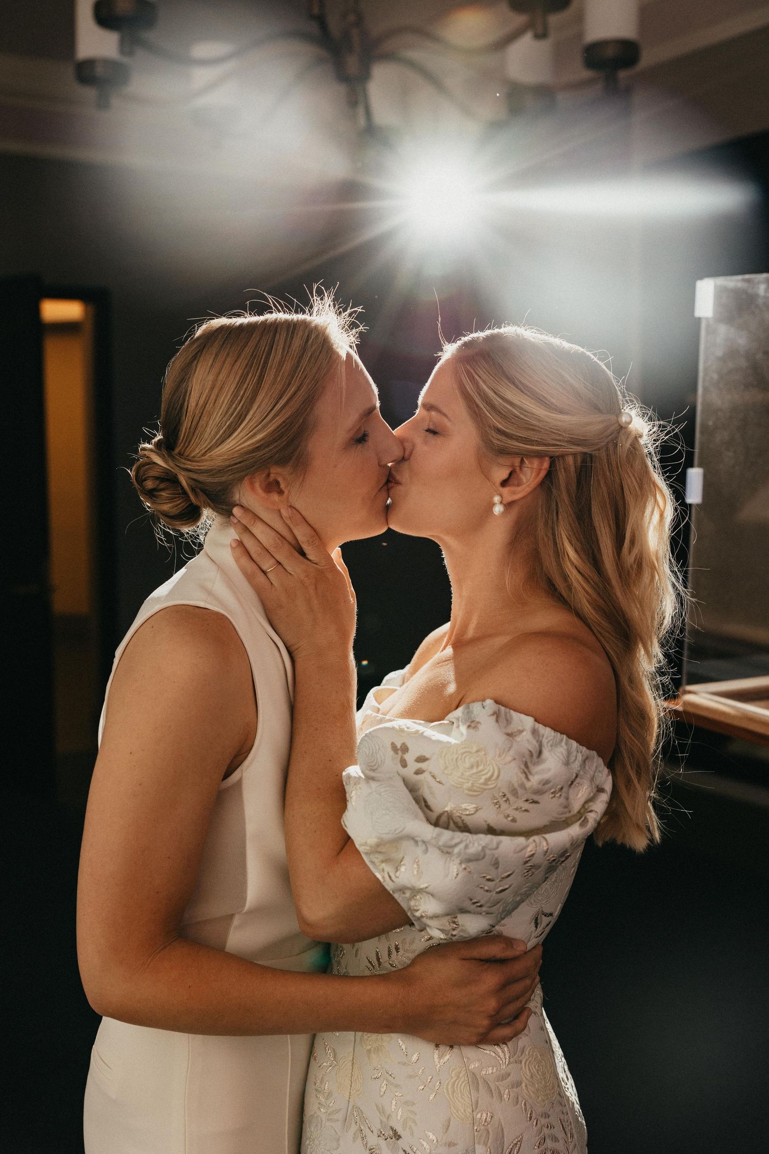 Two brides sharing a romantic kiss in dramatic backlit lighting at their wedding reception.
