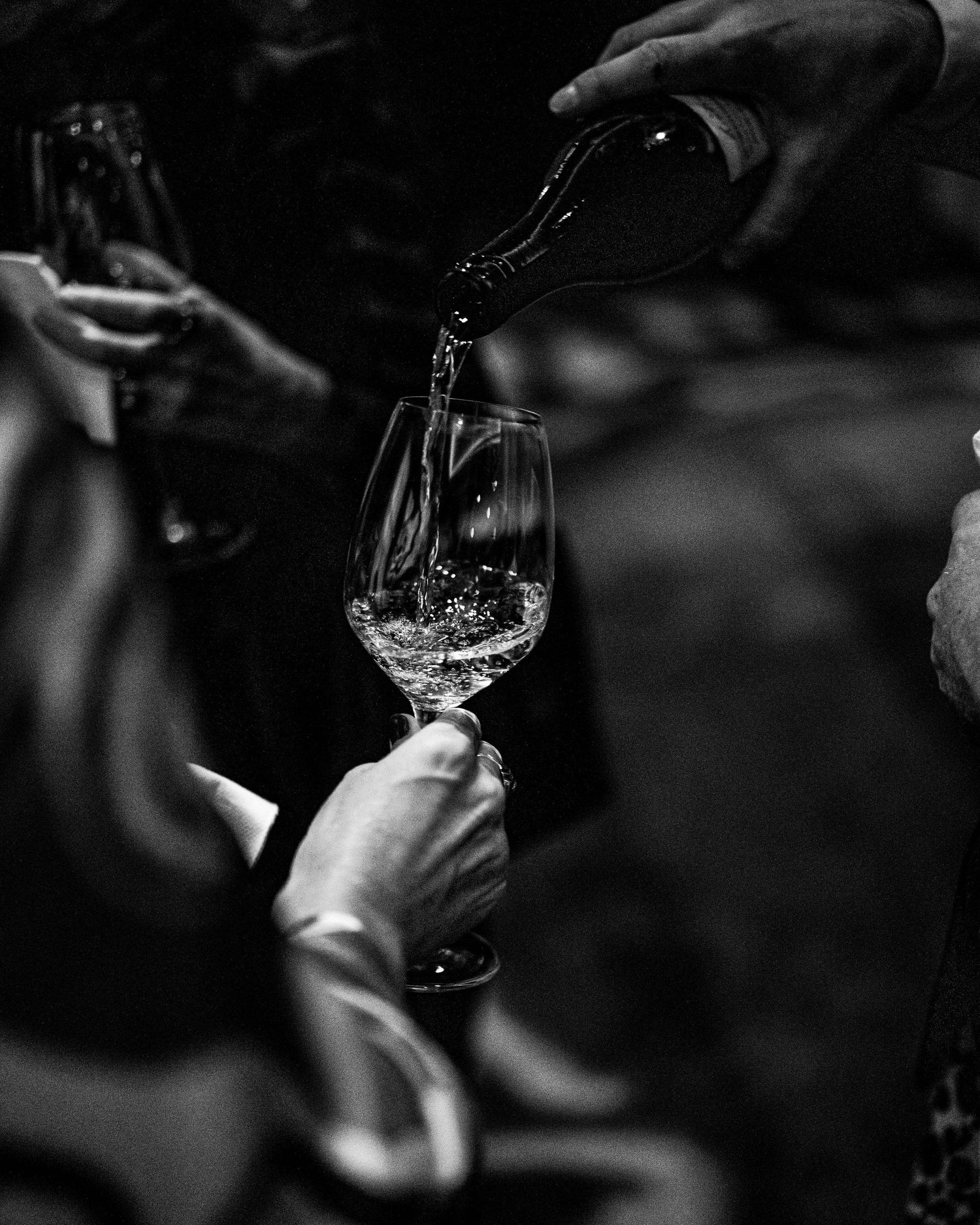 Black and white photo of a hand pouring white wine from a bottle into a glass held by a guest at an elegant tasting event.