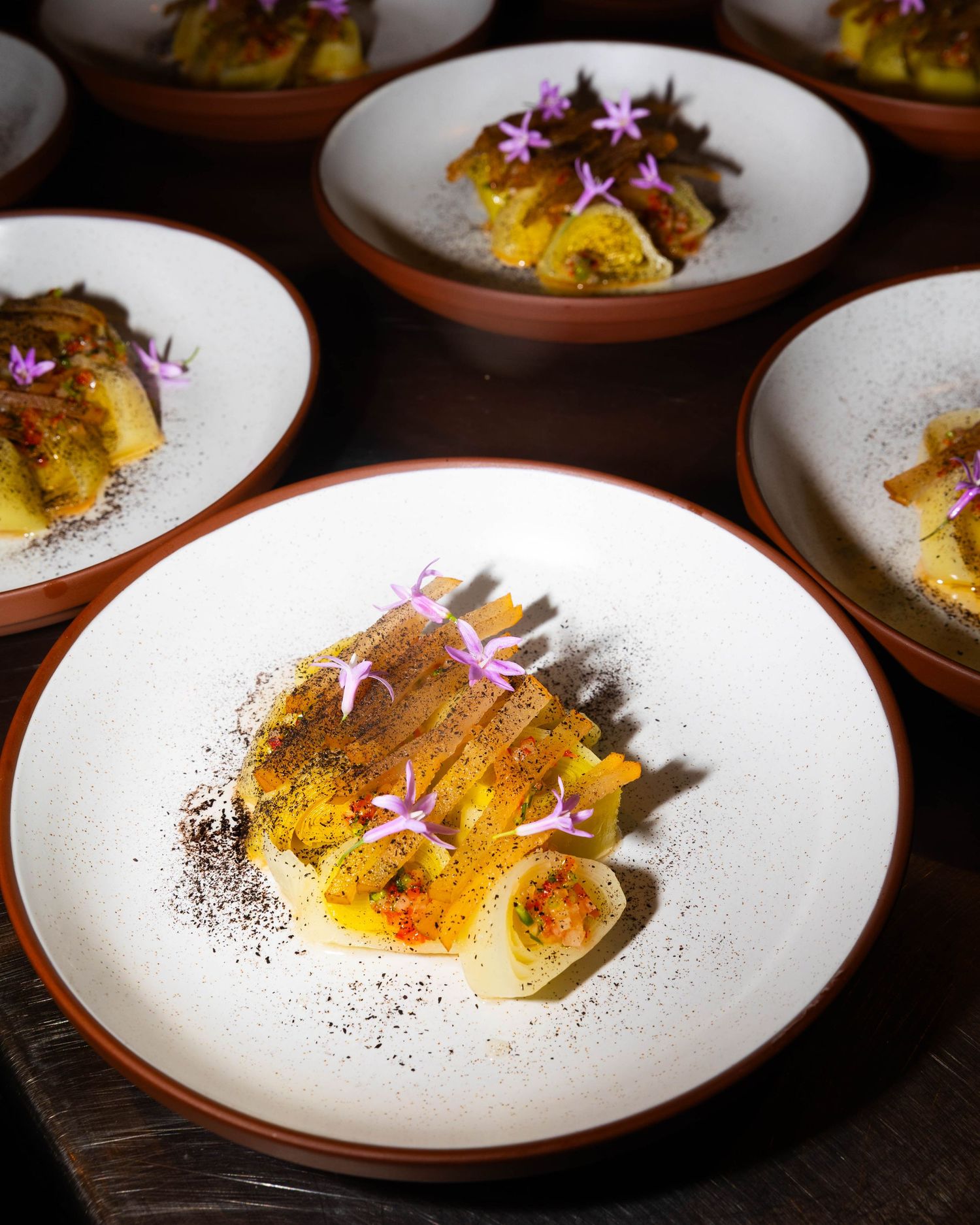 Rustic terracotta-rimmed plates of artfully plated vegetable dishes garnished with delicate pink flowers on a dark wooden table.