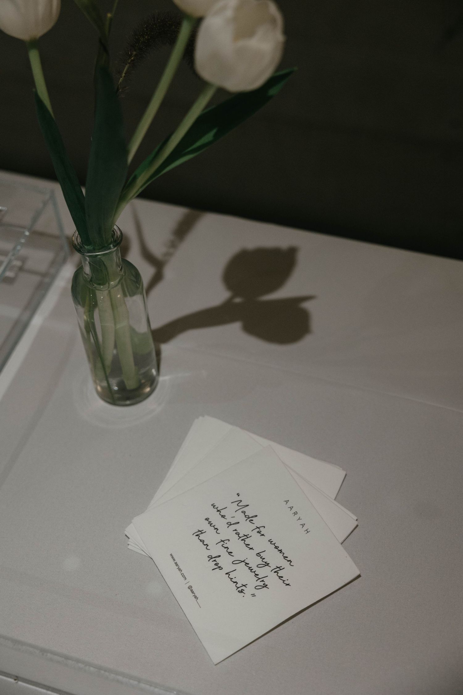 White tulips in a glass bud vase casting a shadow beside handwritten quote cocktail napkins on a white draped table.