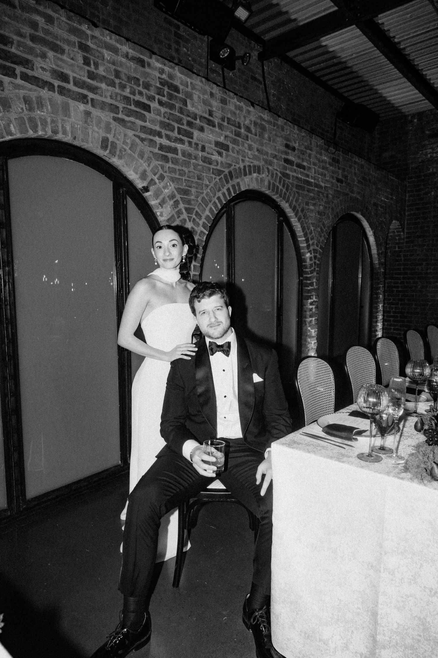 Black and white photo of bride and groom at wedding reception table in brick venue.