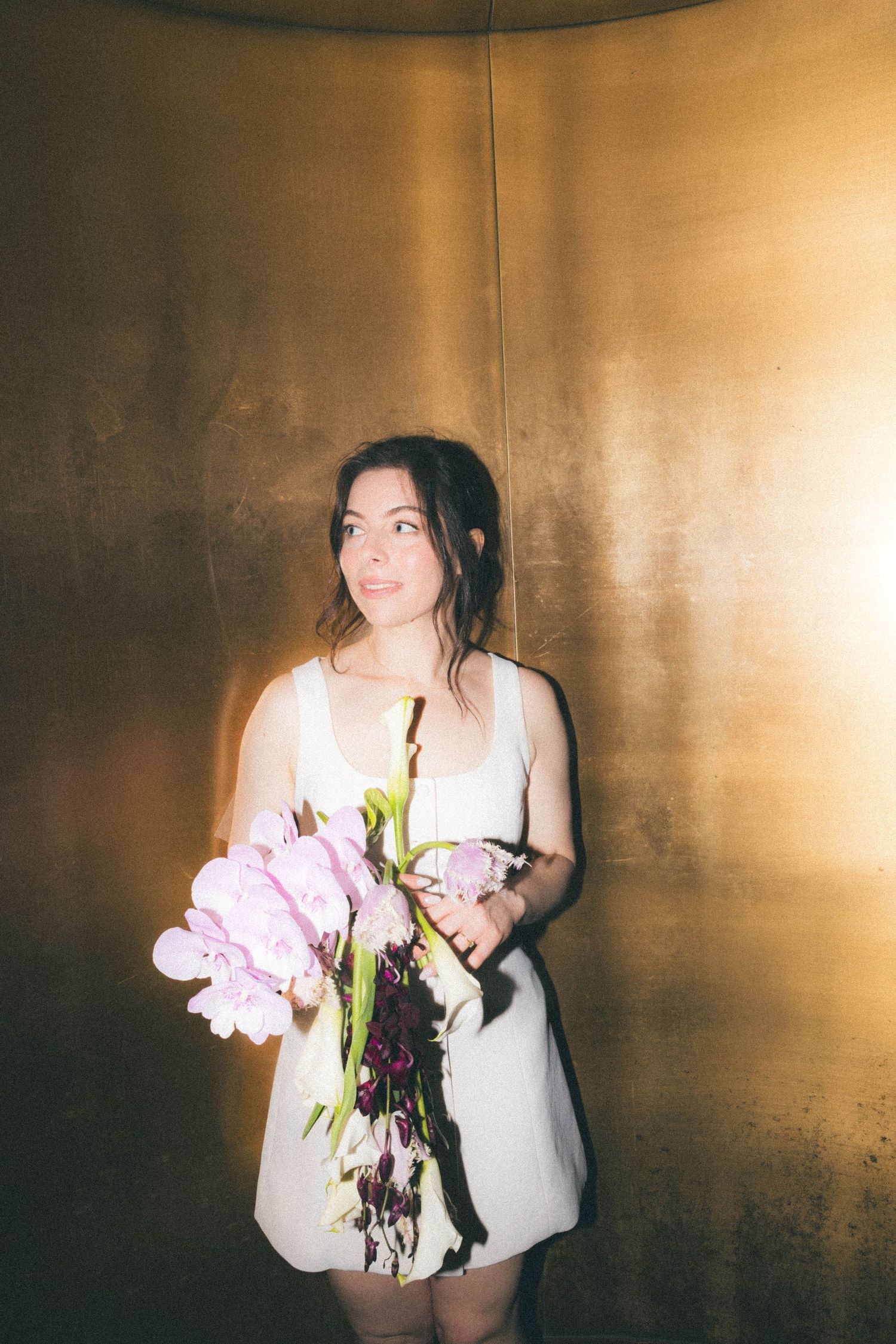 Bride in a white mini dress holding pink peonies bouquet against a golden metallic wall.