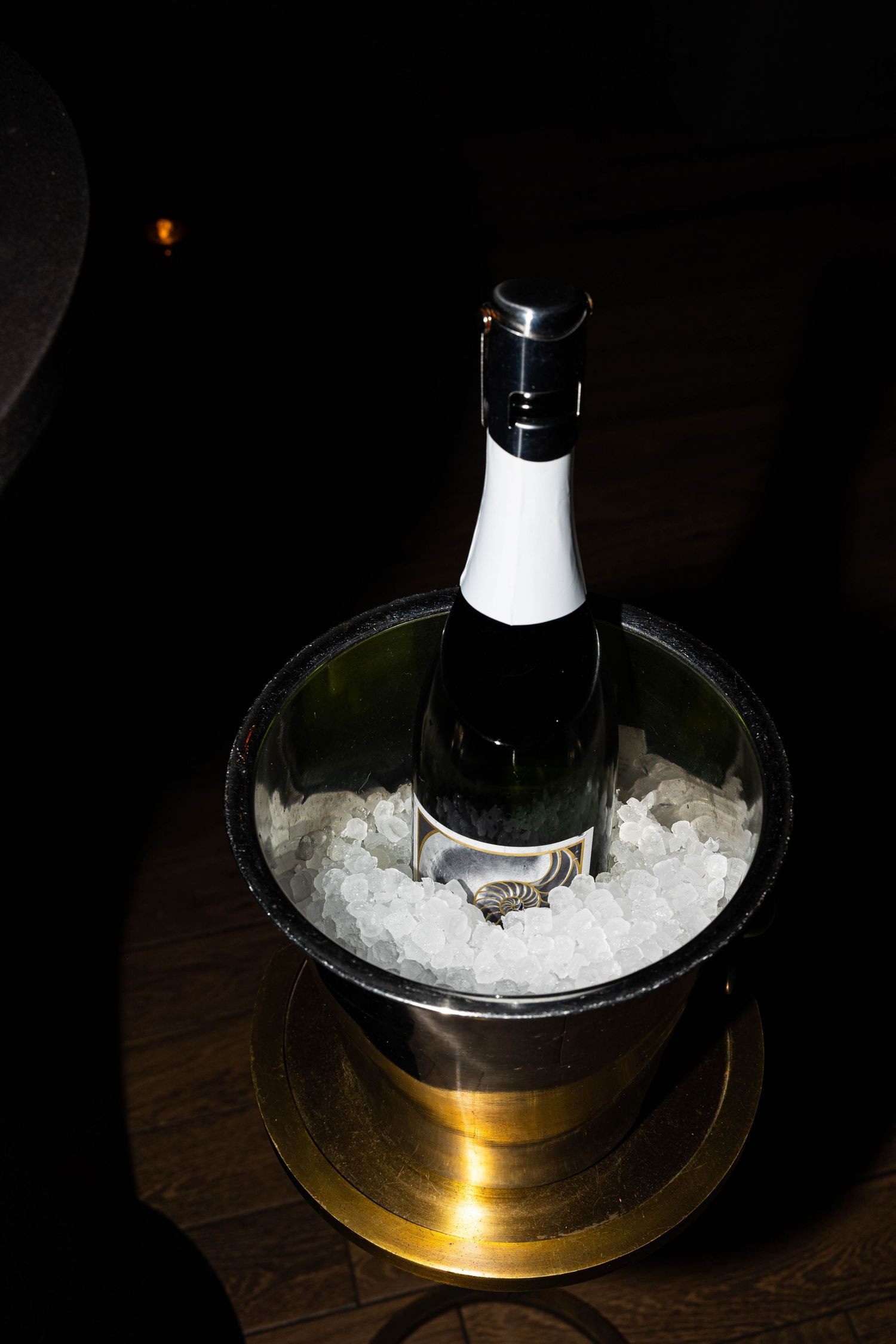 Champagne bottle chilling in ice-filled silver bucket on a gold stand in dark setting.