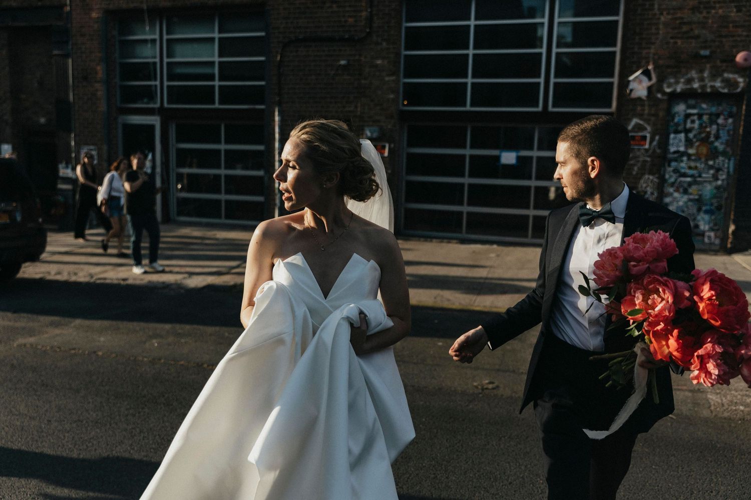 Bride in white gown and groom carrying pink peony bouquet walk together on urban street at golden hour.