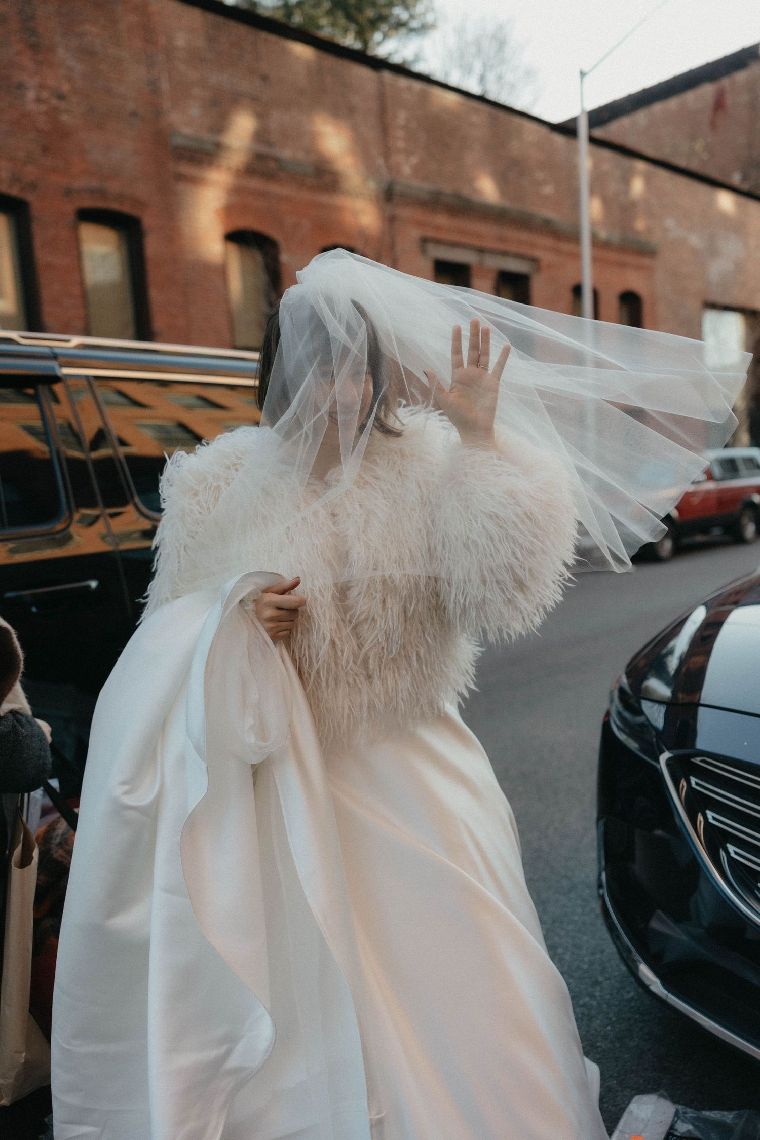 Bride in white gown with feather wrap and flowing veil caught by wind on urban street.
