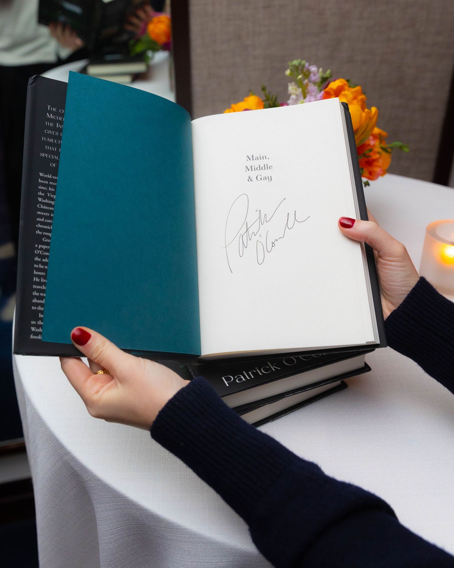 Hands holding an open signed copy of the book Maids, Middle & Gog beside orange flowers and a candle on a white tablecloth.