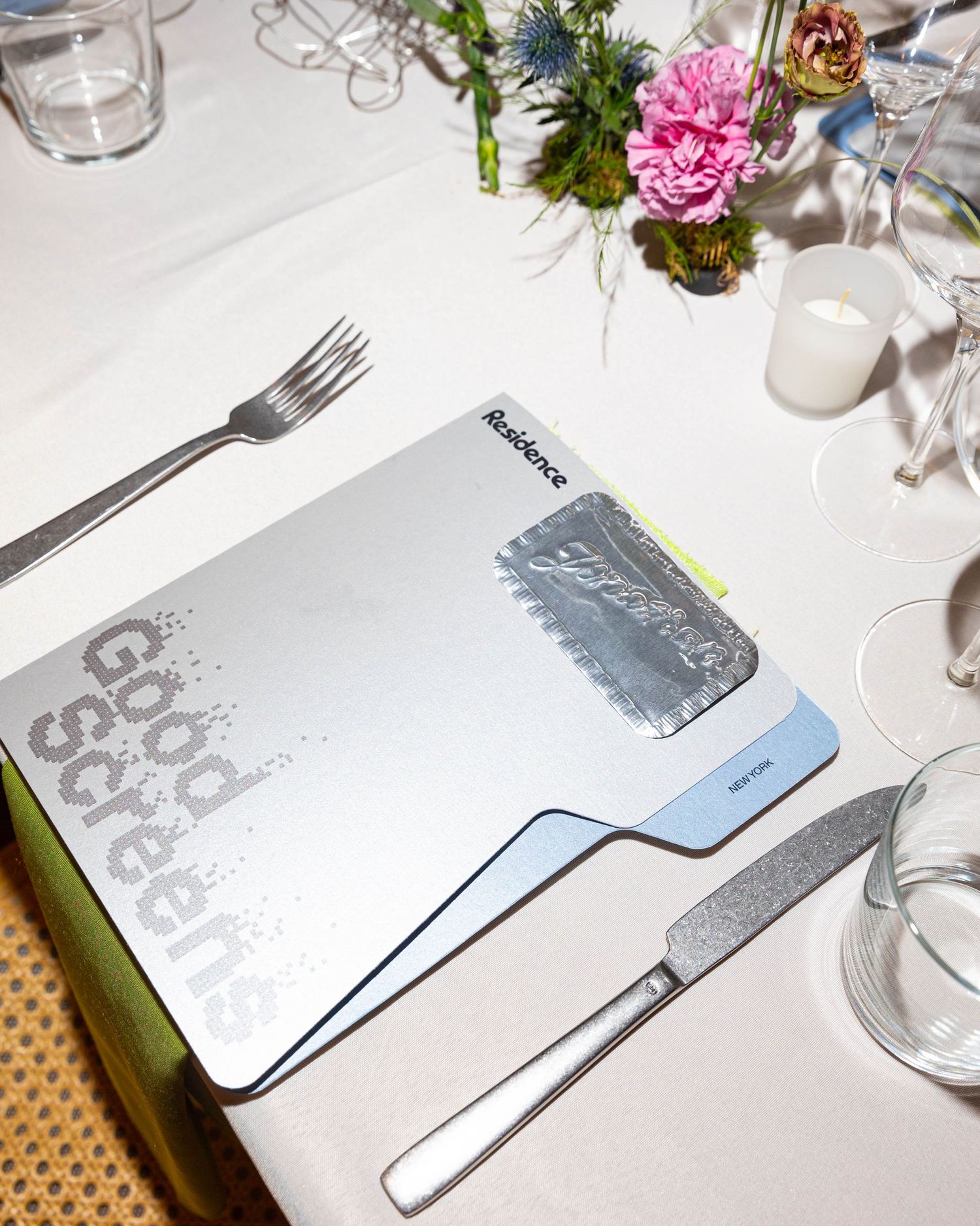 Silver Good Screens event program with a metallic foil panel on a white table beside a fork, knife, glassware and pink flowers.
