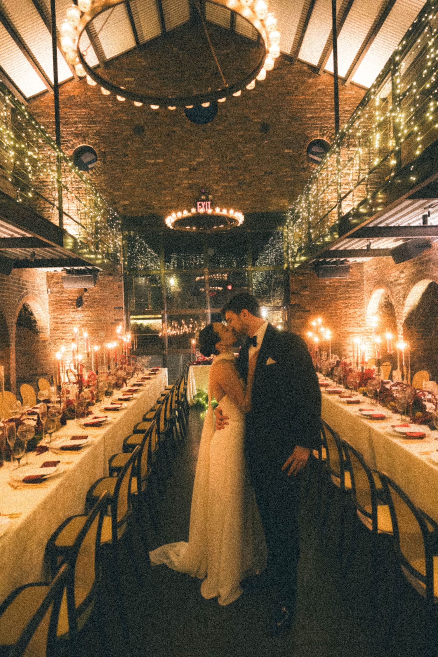 Bride and groom sharing romantic kiss in stunning brick wedding venue adorned with fairy lights.