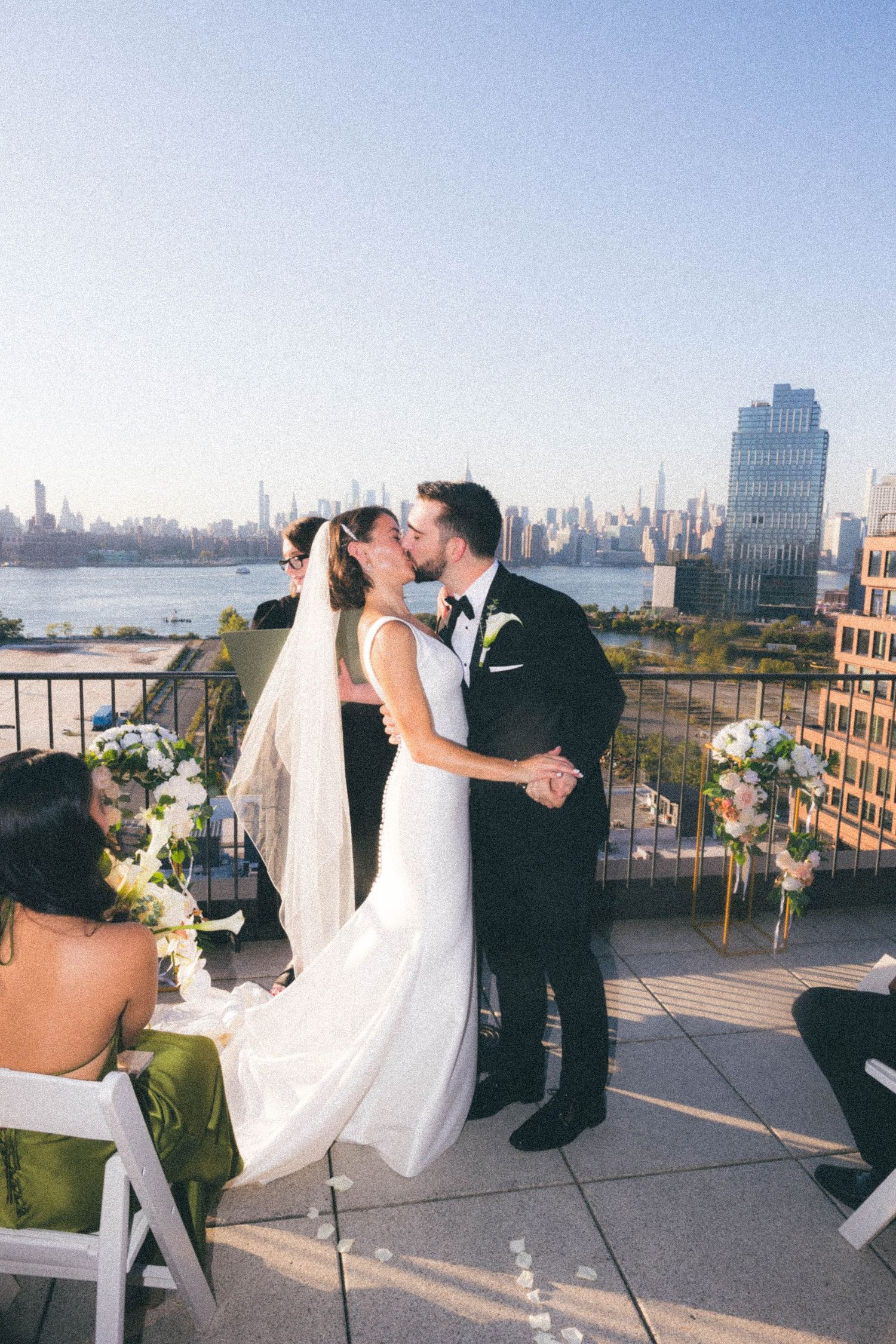 Bride and groom sharing first kiss on rooftop ceremony with NYC skyline and East River backdrop.
