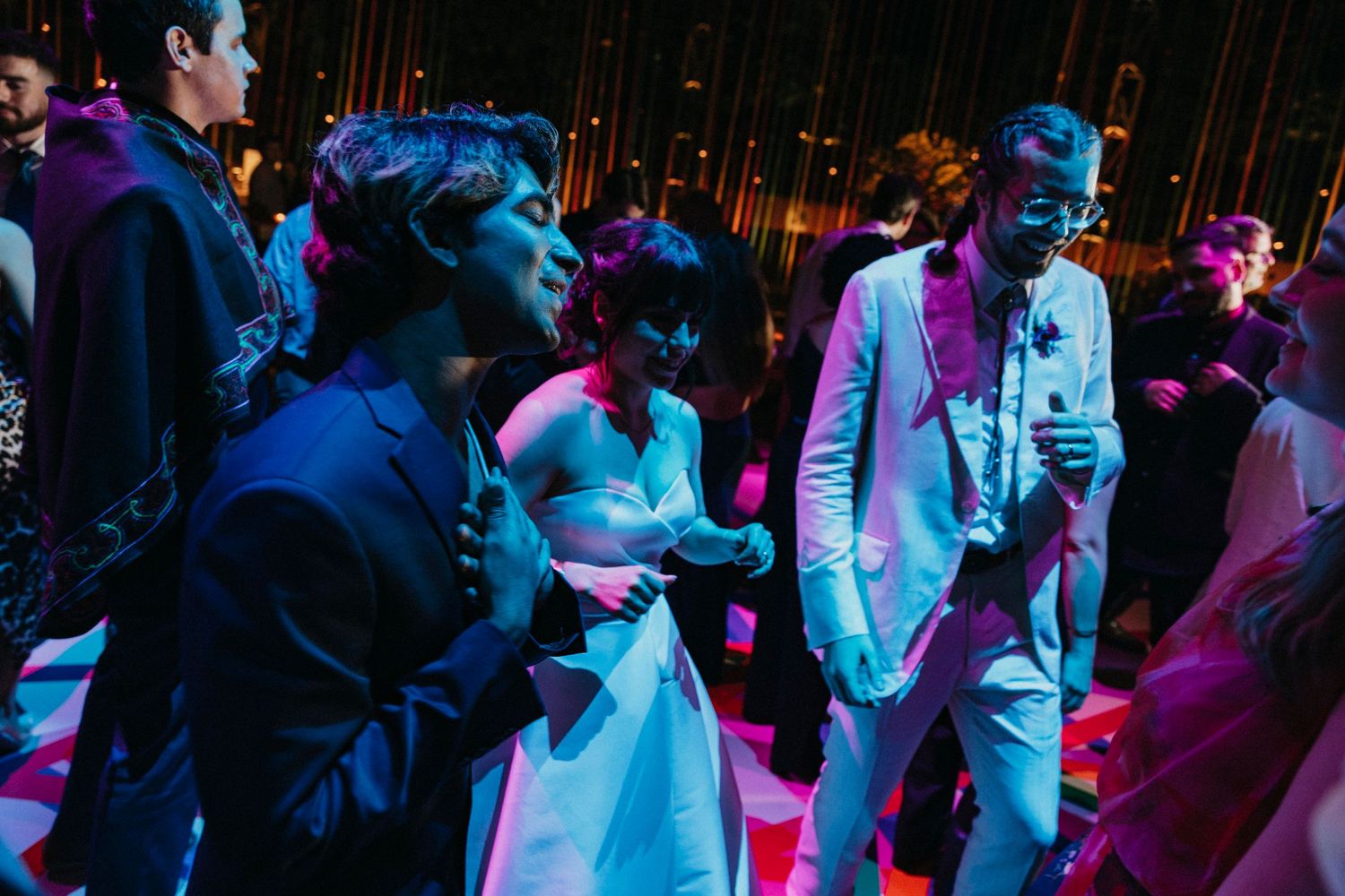 Wedding guests dance joyfully on a colorfully lit dance floor during a lively reception celebration.