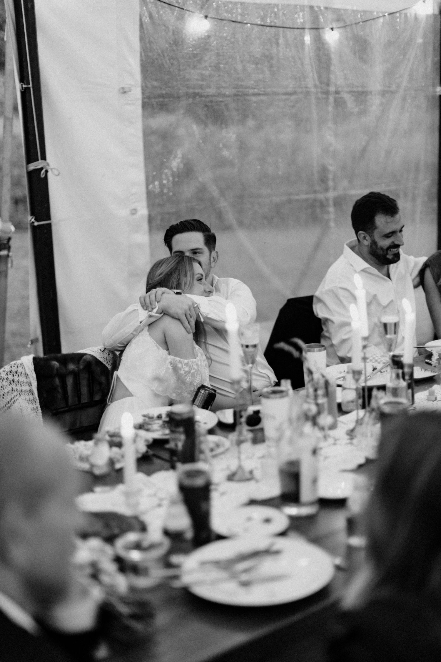 Black and white photo of couple sharing an emotional hug at wedding reception dinner table.