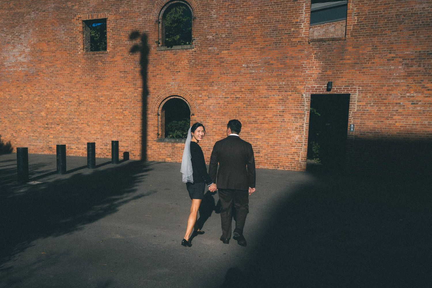 Wedding couple walks hand in hand past rustic brick building in dramatic shadow light.