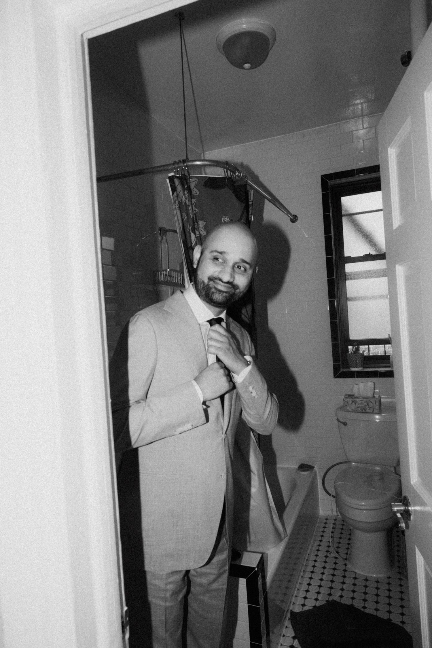 Groom in light gray suit adjusts tie in bathroom mirror in black and white photo.