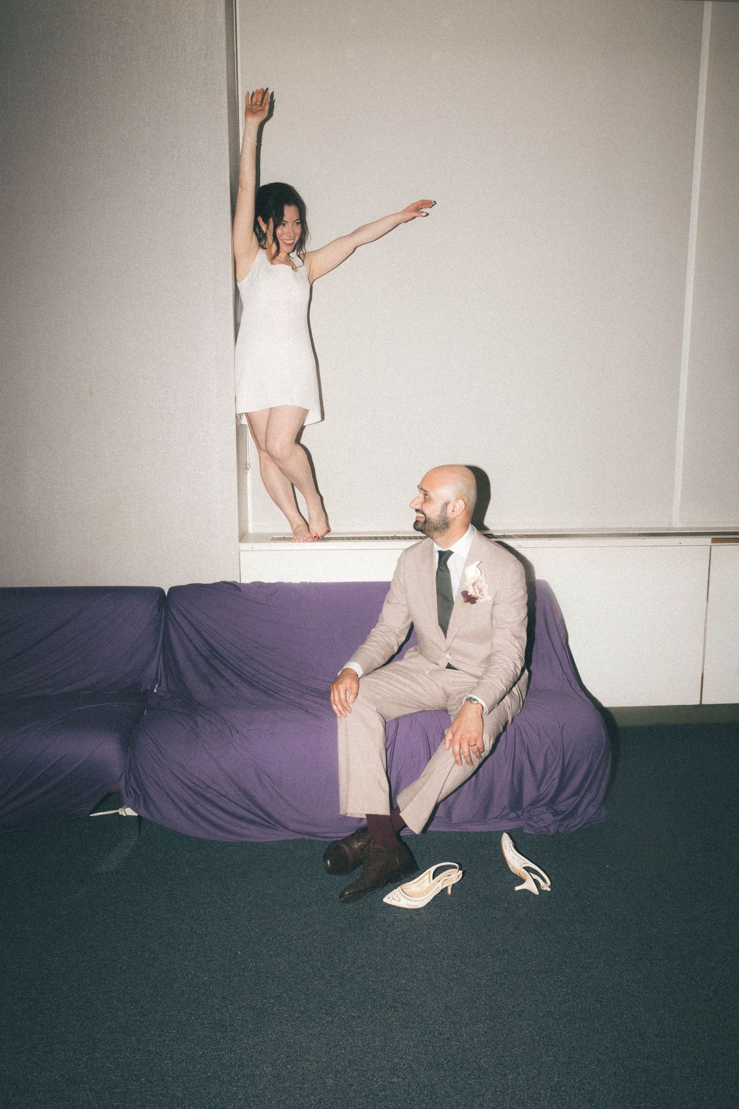 Groom sitting on purple couch watching bride standing on ledge with arms outstretched in joy.