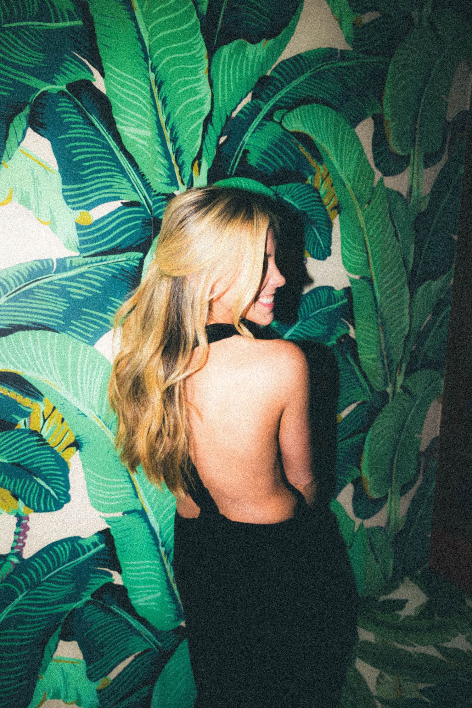 Blonde woman in backless black dress smiling against tropical banana leaf wallpaper.