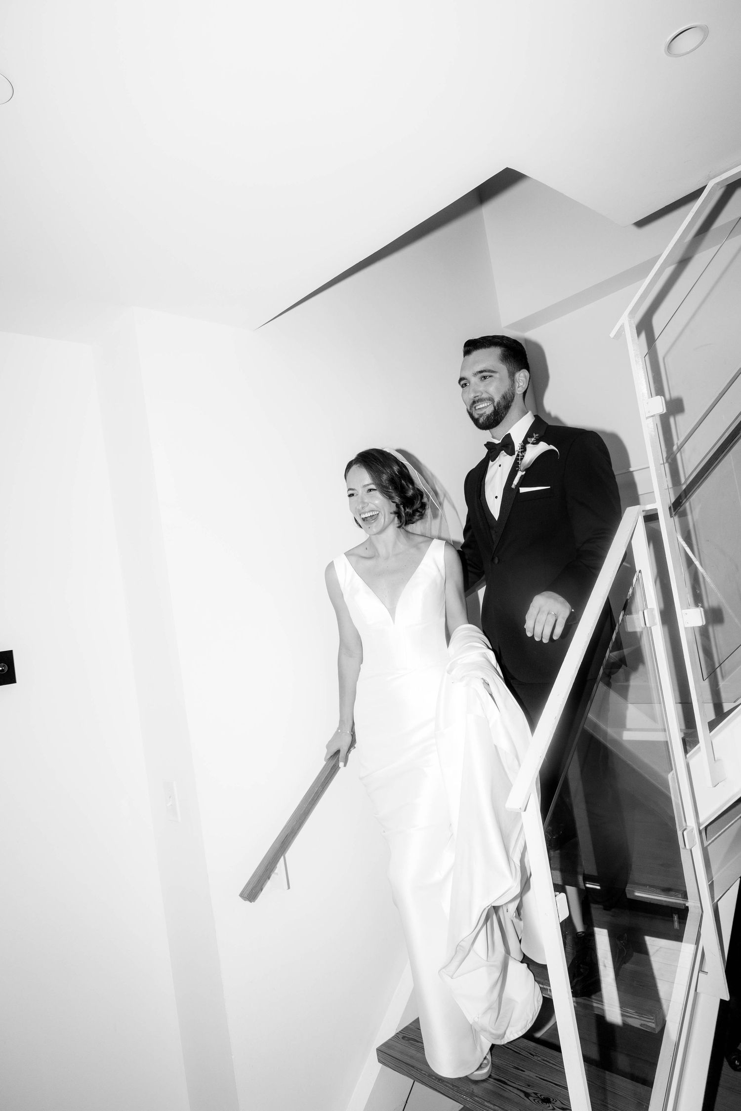 Laughing bride in elegant white gown and groom in tuxedo descending modern staircase in black and white.